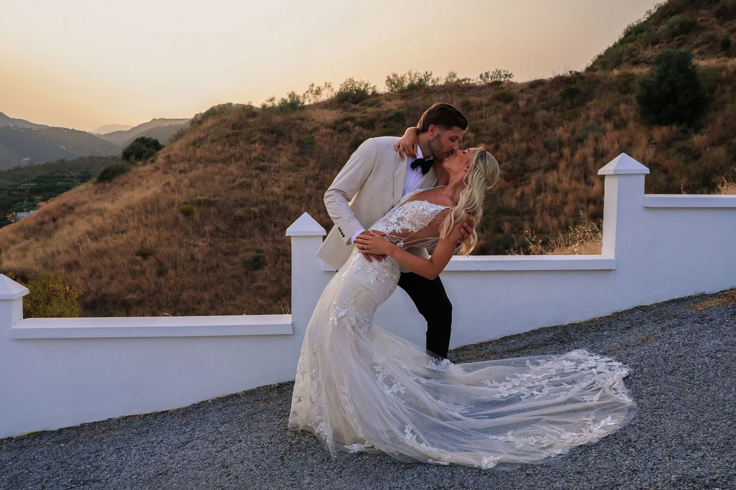 Couple sharing a romantic dip kiss at sunset, bride in lace gown and groom in white suit, with scenic hills backdrop.