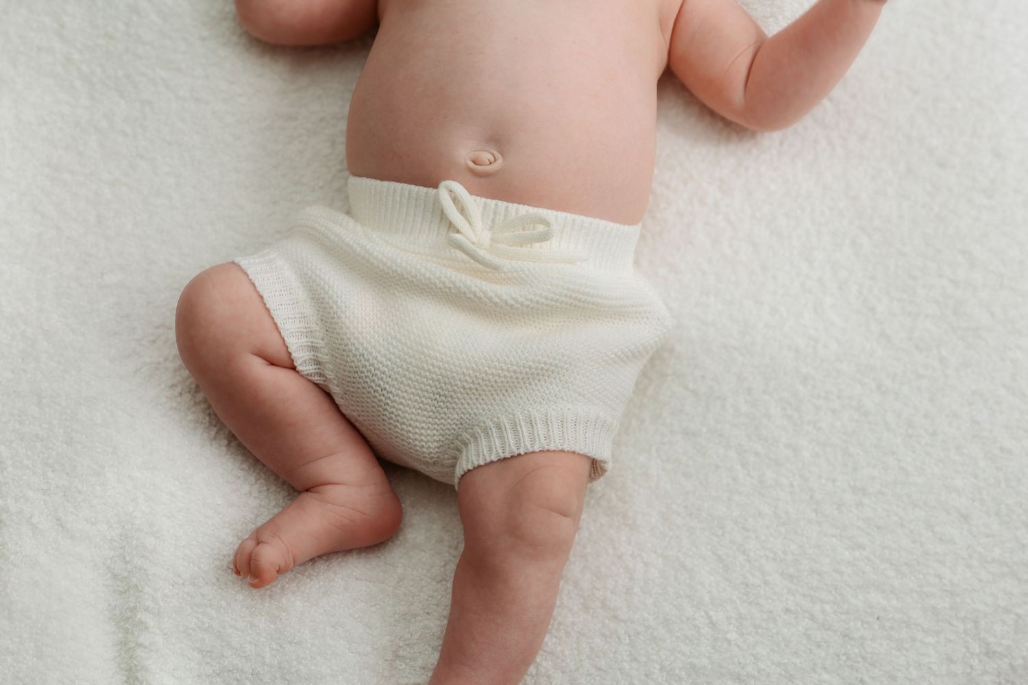 Baby lying on white blanket wearing cream knit bloomers shorts, showing chubby legs and belly button.