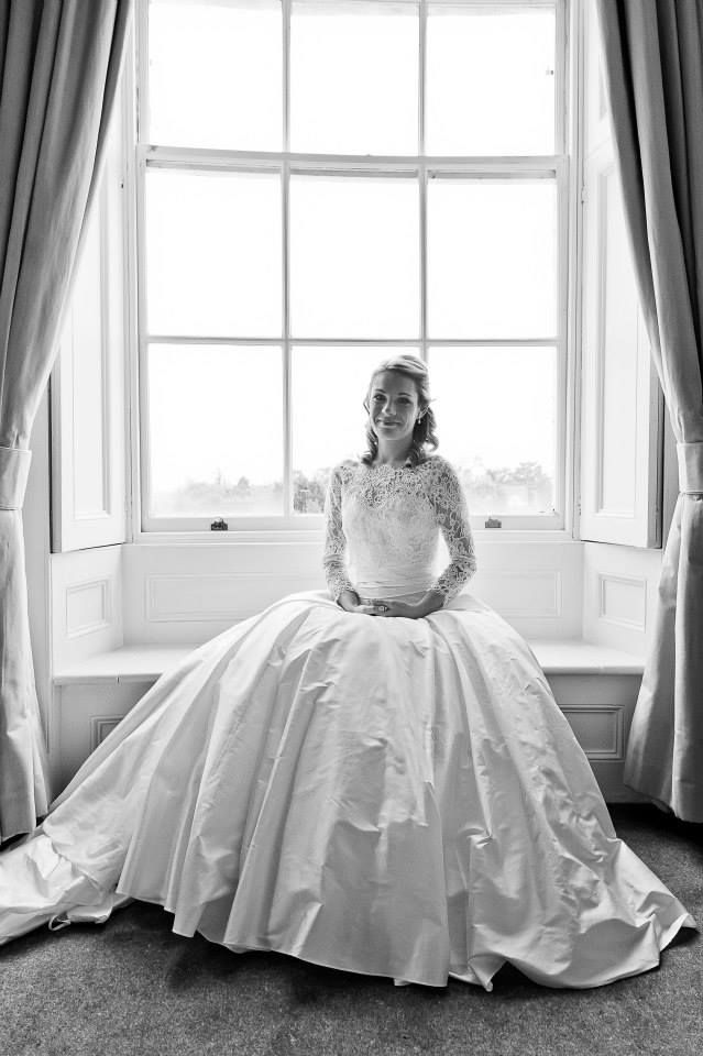 Elegant bride in lace-detailed ball gown seated by large window in black and white.