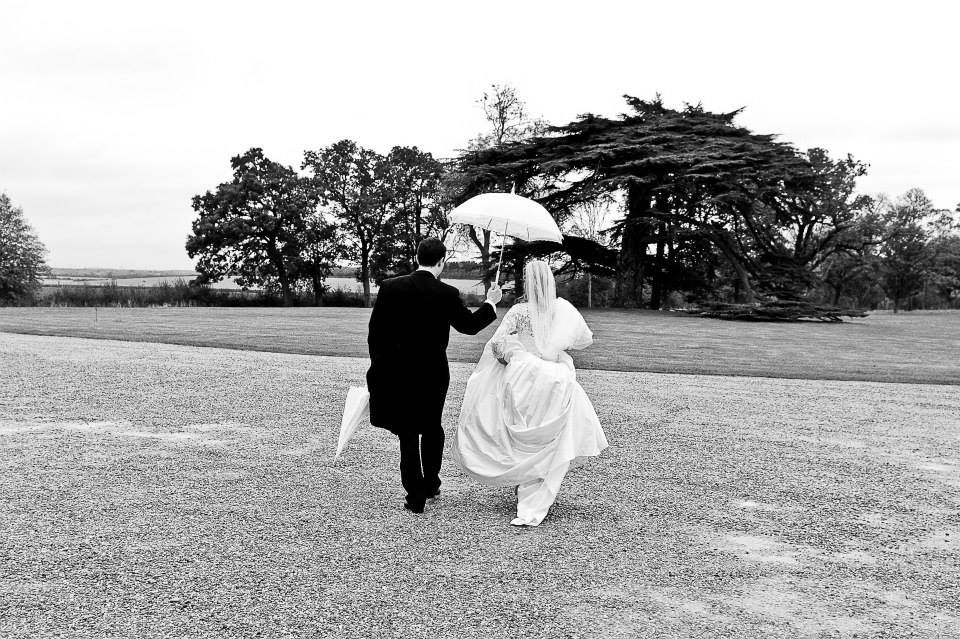 Black and white photo of couple running across field with umbrella on wedding day.