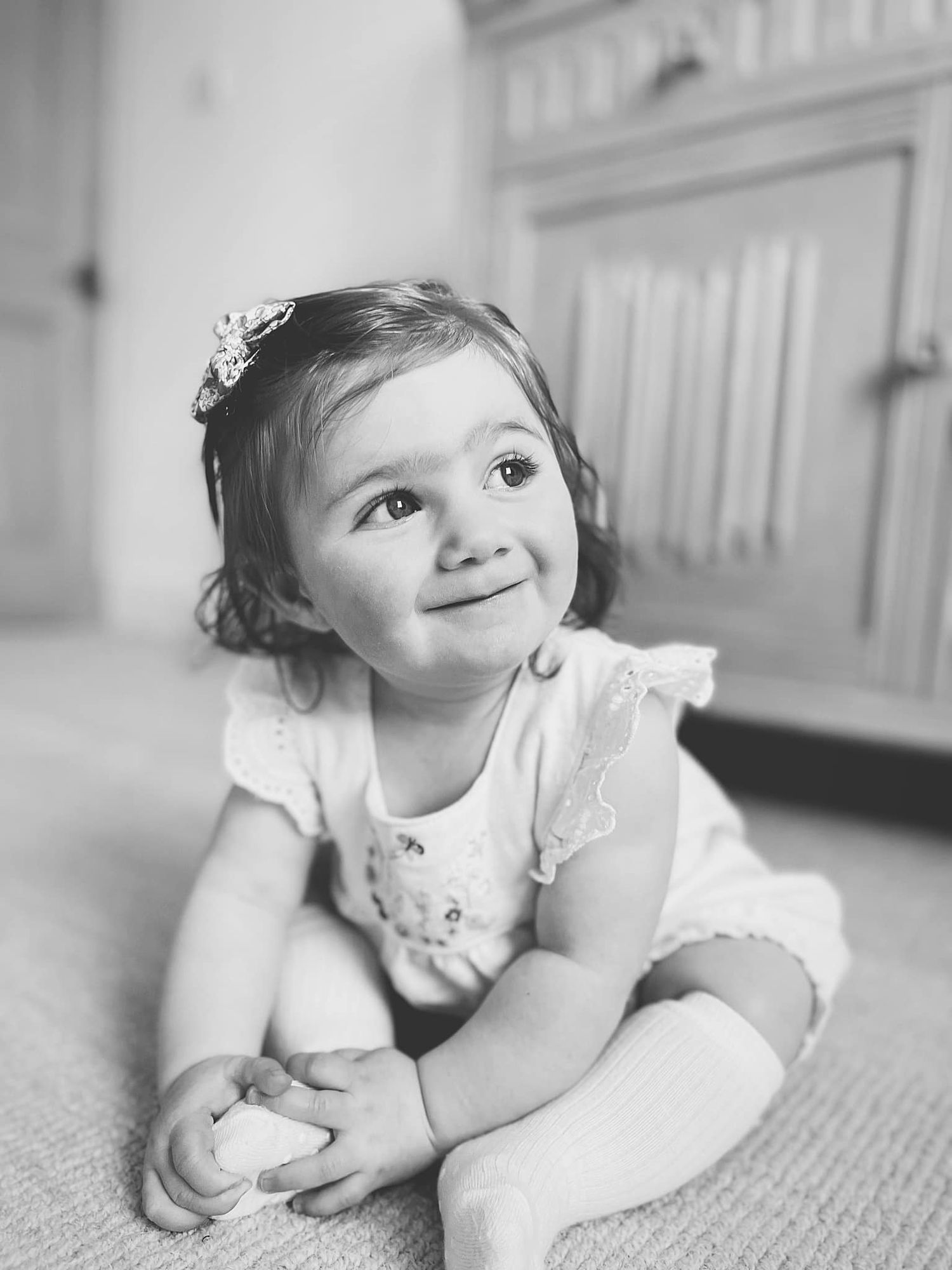 Black and white portrait of a toddler in lace dress lying on a carpet.