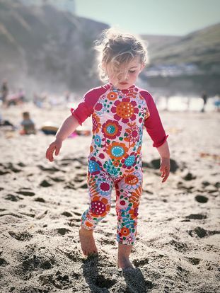 Child wearing colorful floral print swimsuit plays in sand at beach on sunny day.