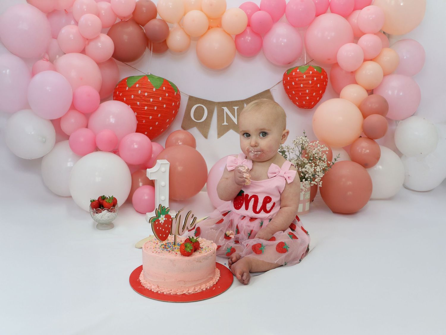 Pink and peach balloon arch decorates a first birthday party setting with cake and strawberry themed decor.