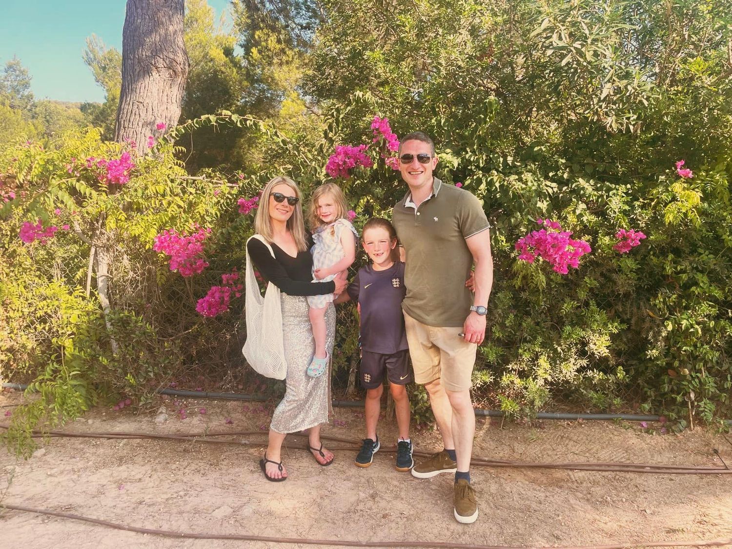 Family of four posing together in front of flowering bushes on a sunny day.