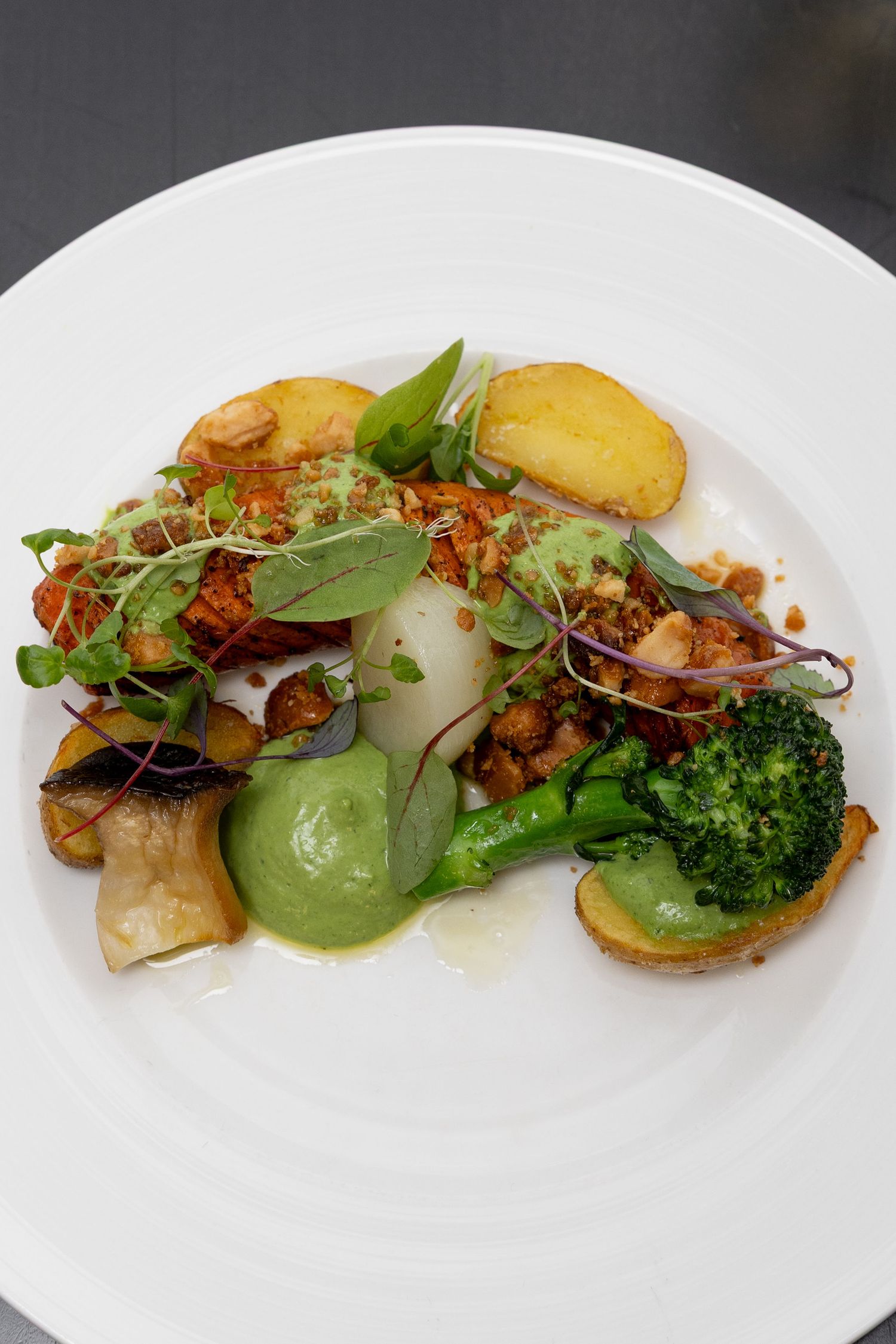 Modern plating of roasted vegetables with green puree, herbs, and crispy elements on a white plate.