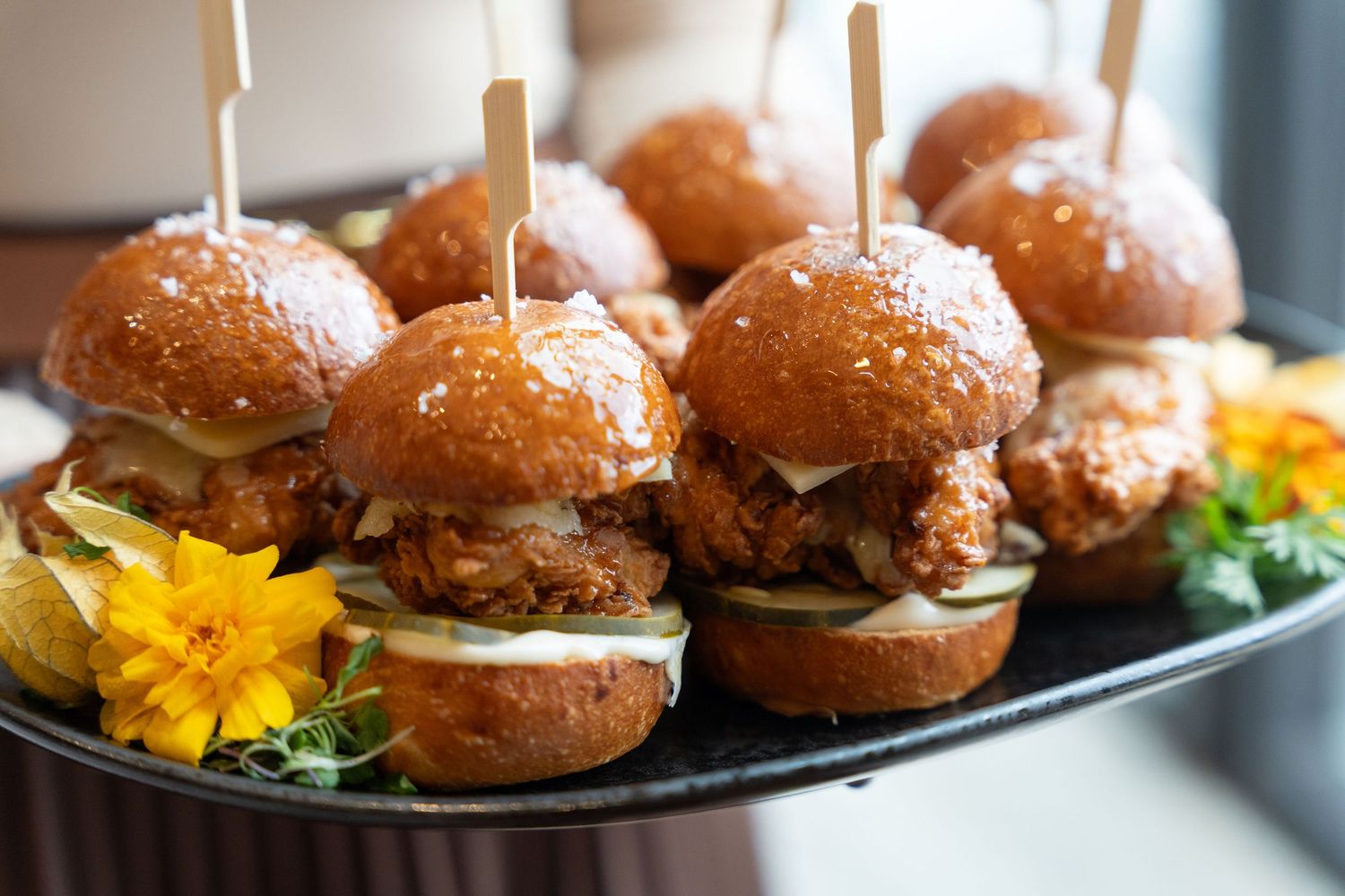 Line of artisanal mini burgers with toothpicks served on a dark platter with decorative flowers.