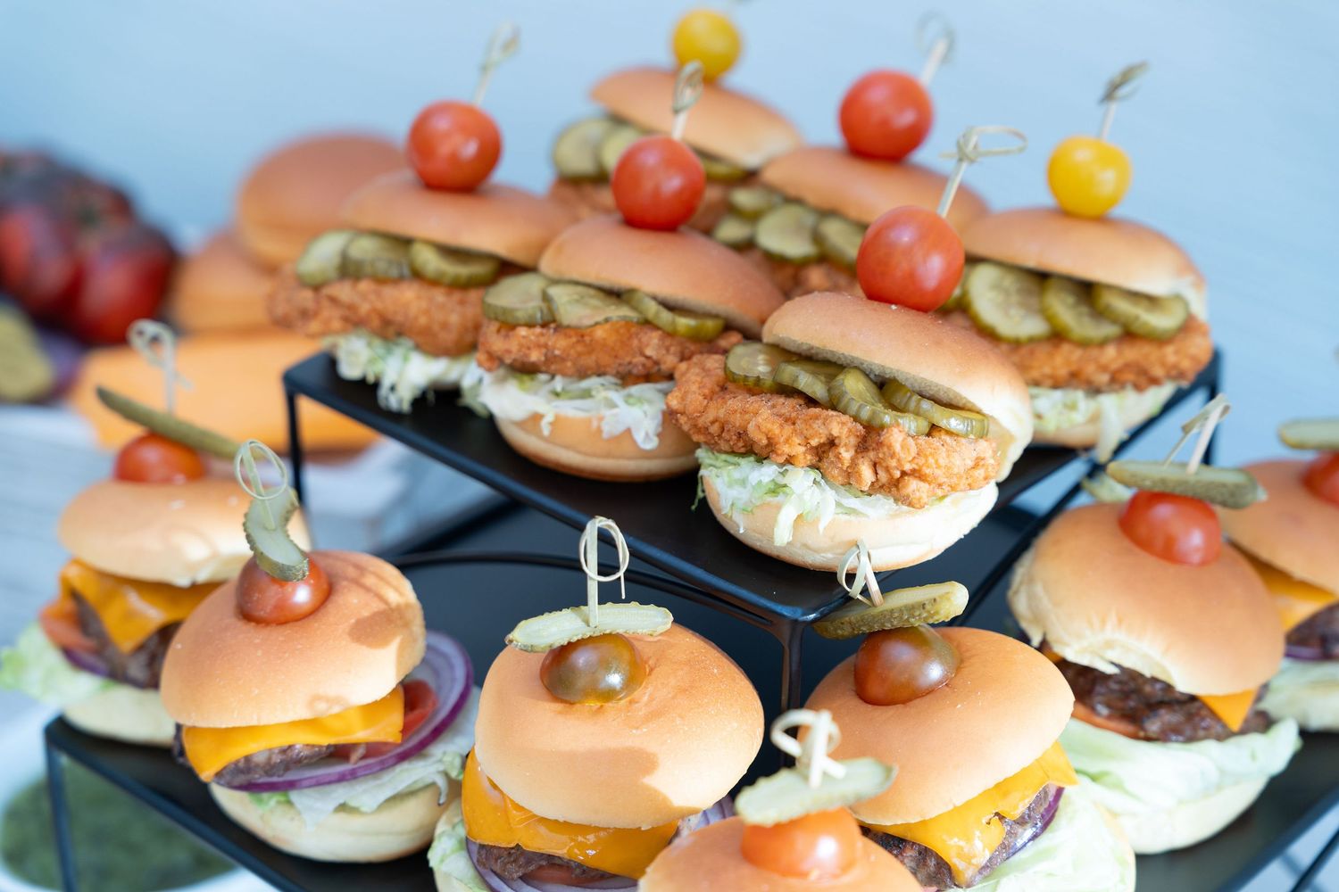Assorted mini burgers and sandwiches arranged on a tiered serving platter with colorful cherry tomatoes.