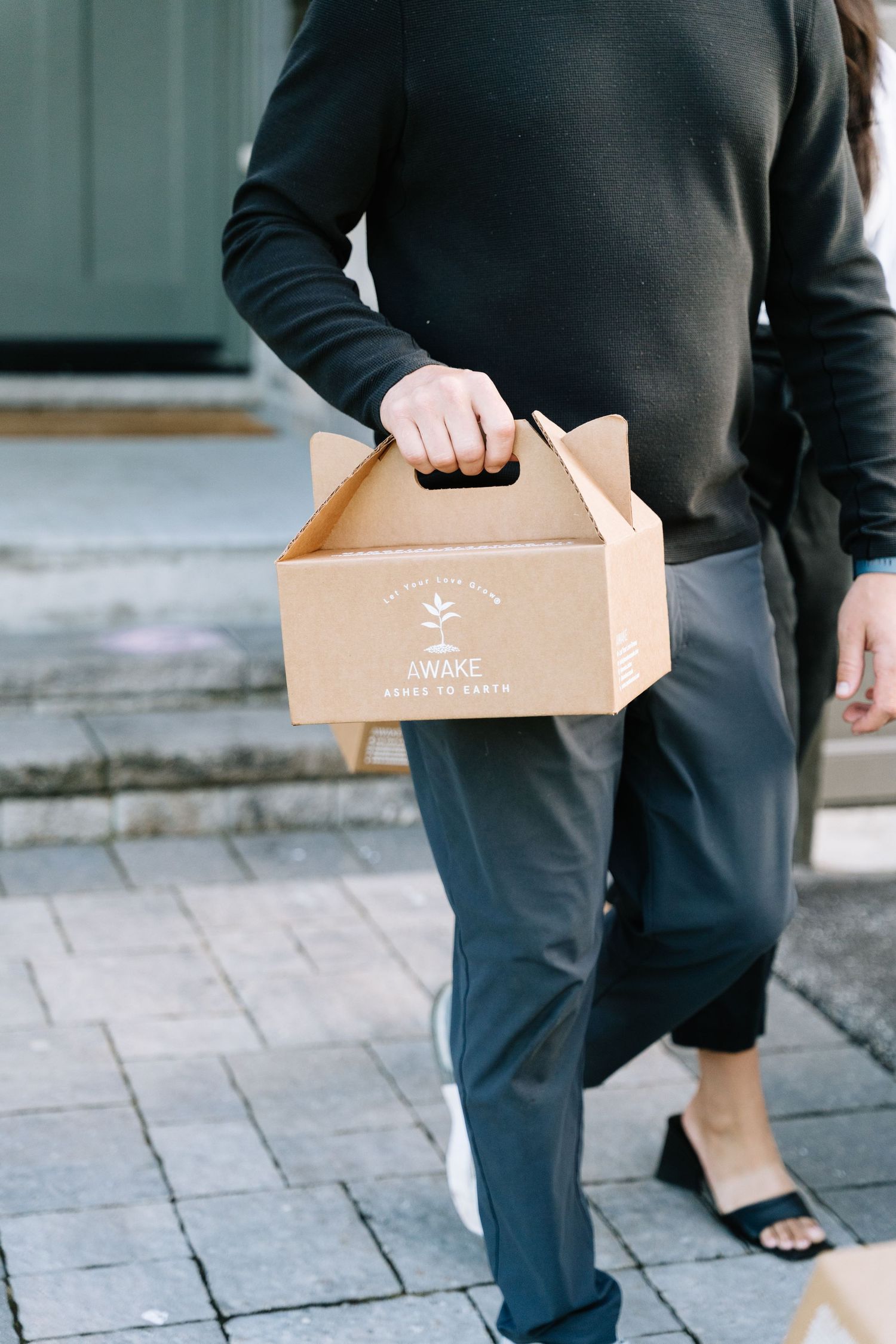 Person in dark clothing carrying a kraft paper takeout box with a handle on stone pavement.