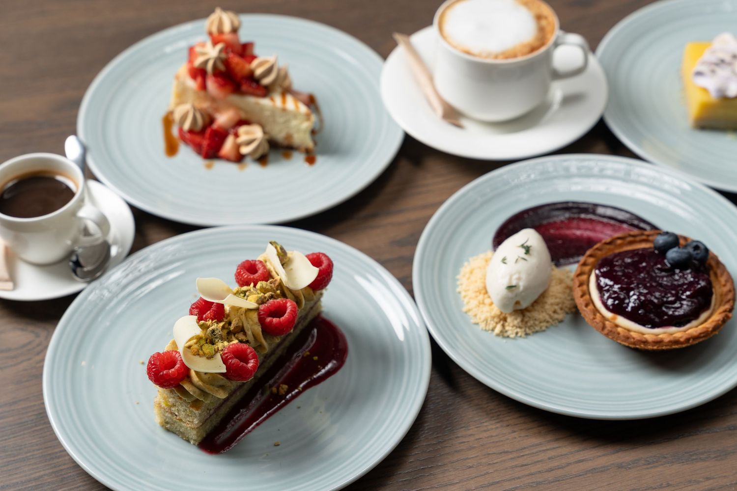 Elegant dessert spread with pastries, tarts, raspberries, and coffee cups on blue plates atop a wooden table.
