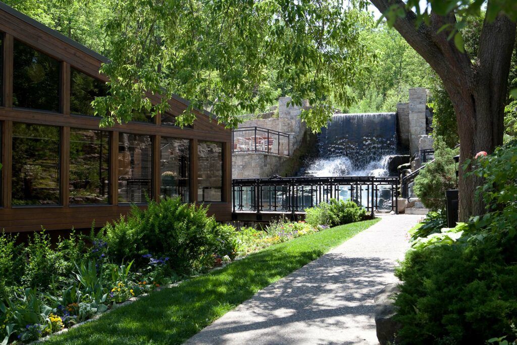 Lush garden pathway leading to a charming brick building with waterfall feature surrounded by mature trees and greenery.