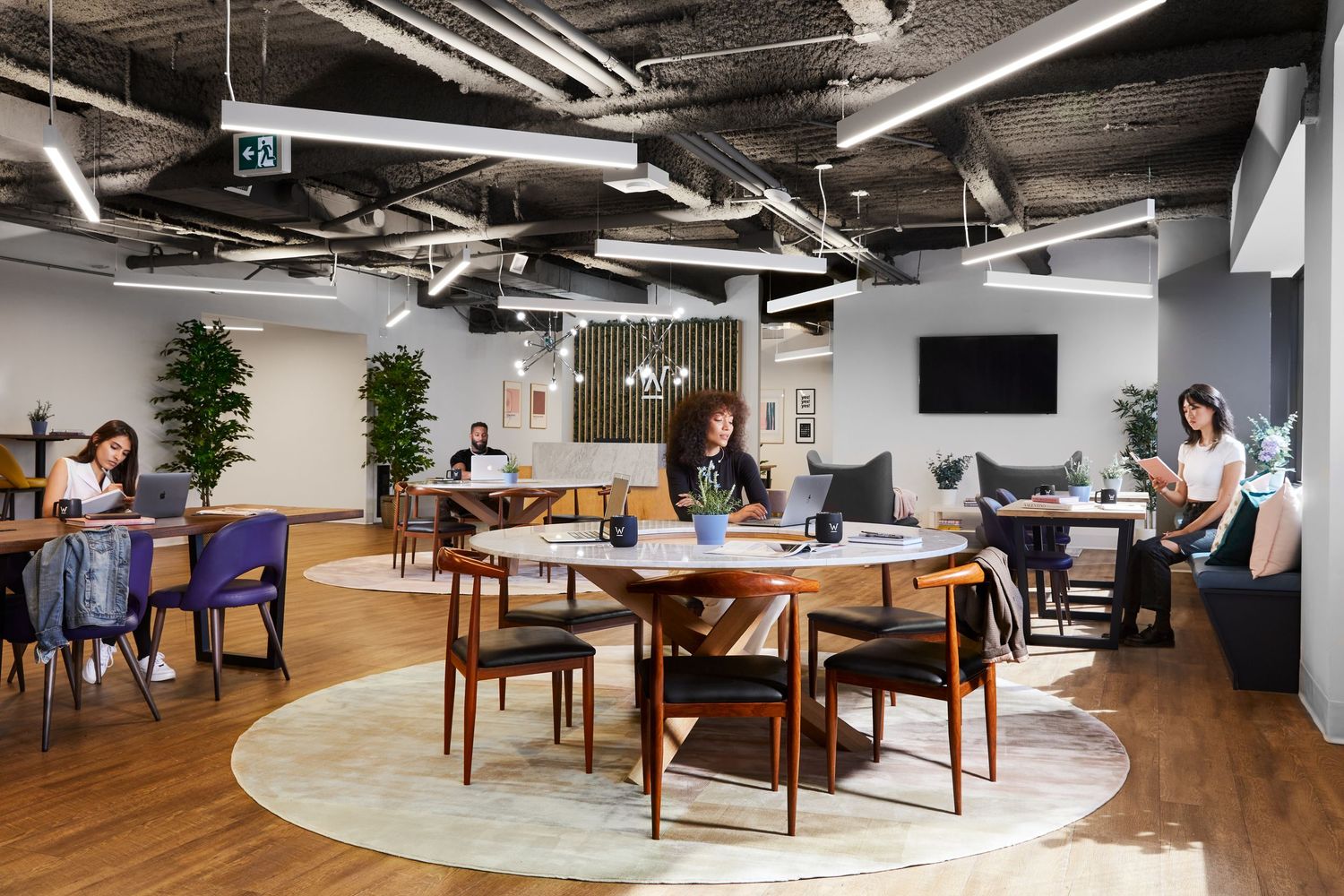 Modern coworking space with round tables on area rugs, wooden floors, and industrial ceiling details.