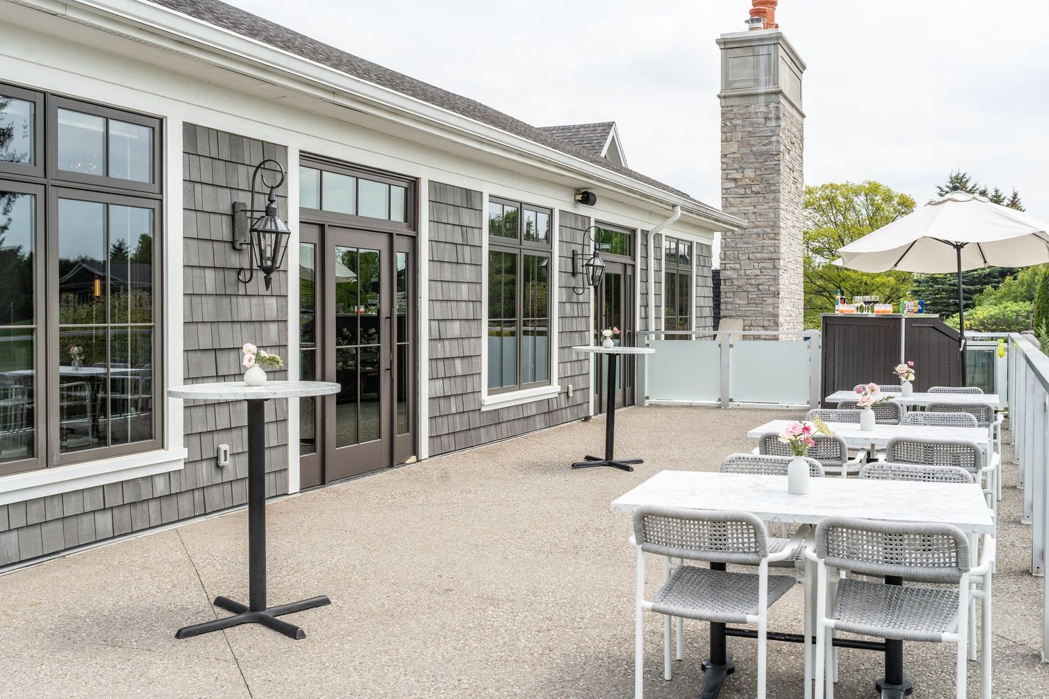 Outdoor patio area of a modern building with grey stone walls, large windows, and white umbrellas for shade.