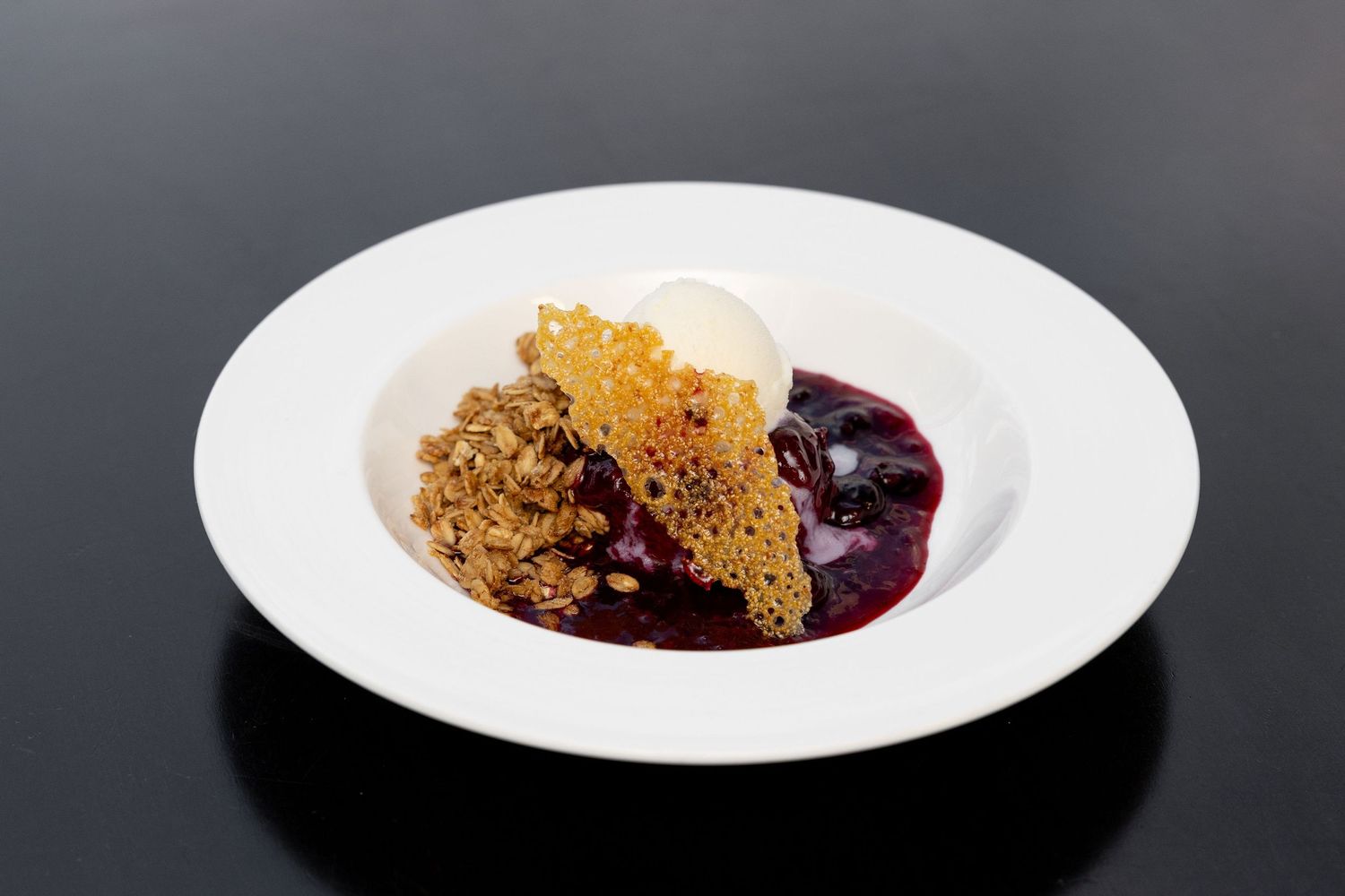A rustic berry crumble dessert with golden brown topping served on a white plate with dark purple sauce.