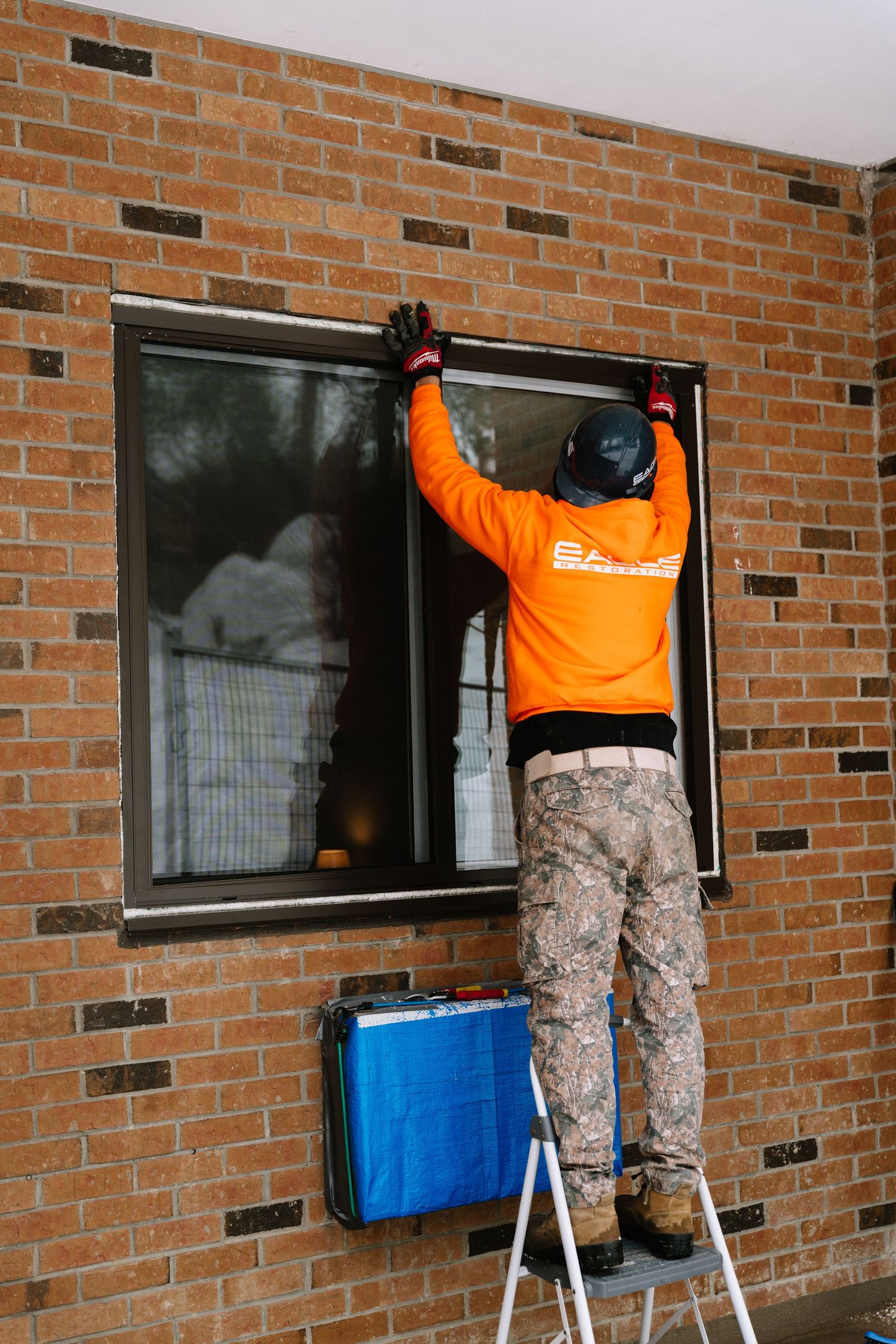Maintenance workers on stepladders clean exterior windows of a brick apartment building.