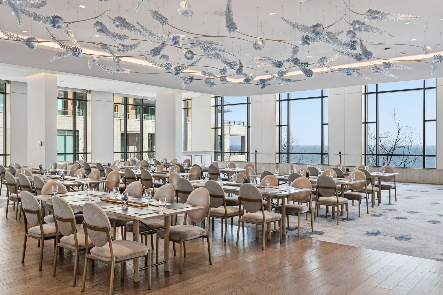 Modern open office space with floor-to-ceiling windows, white tables and grey chairs in a minimalist setting.