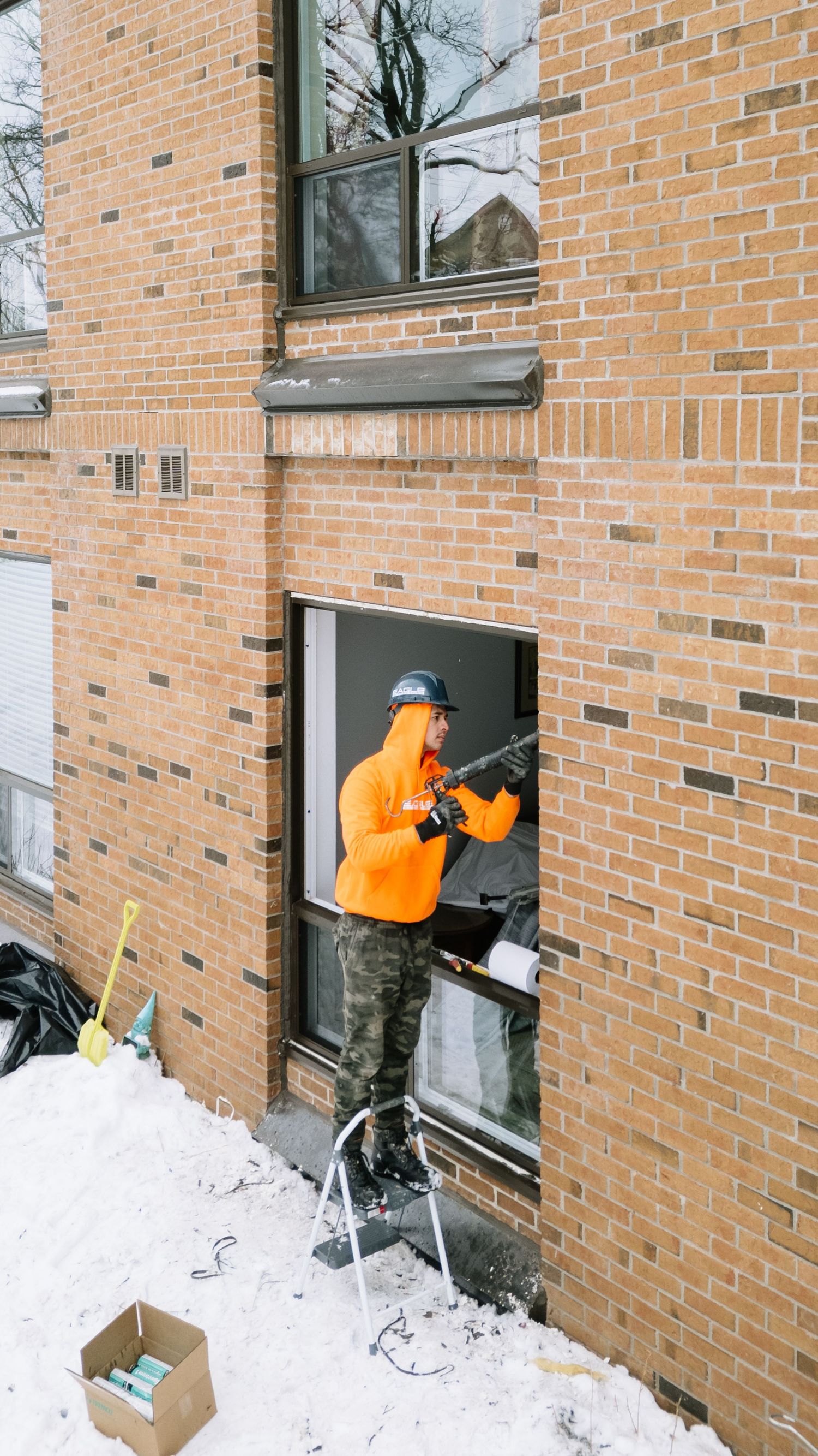 Worker in bright orange jacket clearing snow from window wells of brick building during winter maintenance.