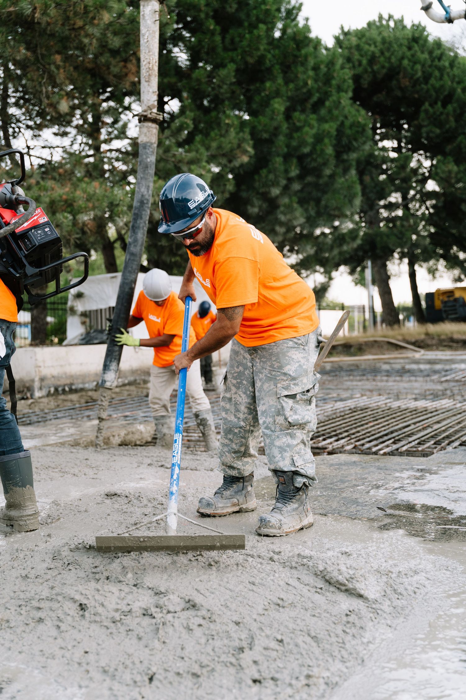 Construction worker in orange safety vest uses power tool to smooth wet concrete surface on outdoor pathway.