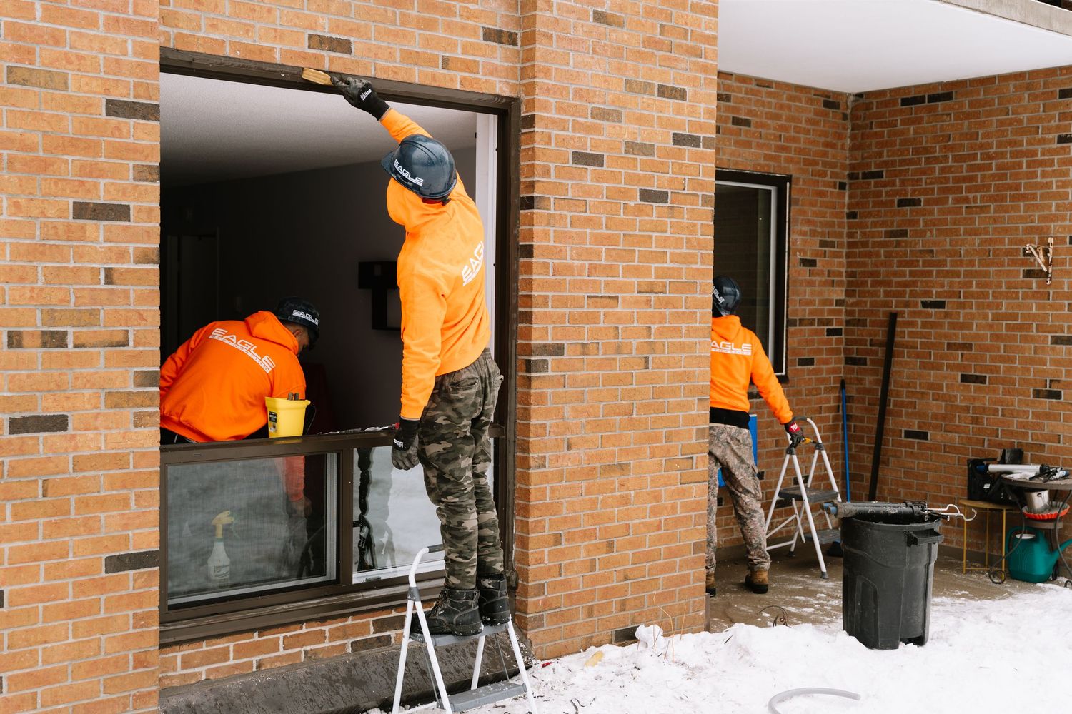 Professional window cleaners work along a row of windows on a brick building exterior.