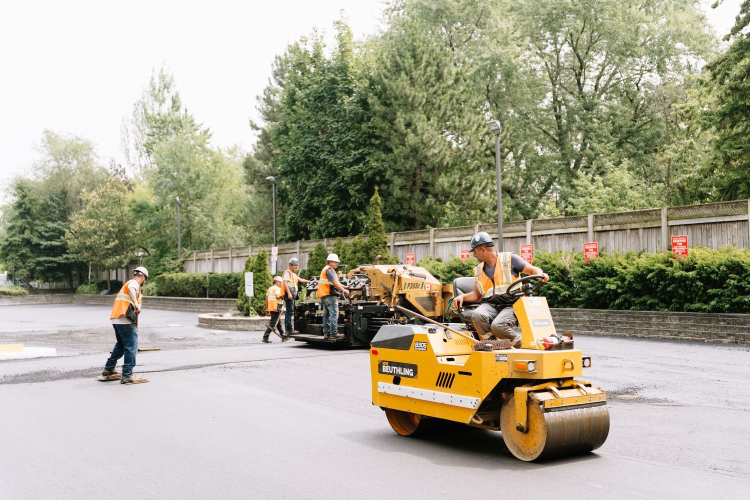 Yellow road roller and paving equipment working on an asphalt surface near trees and buildings.