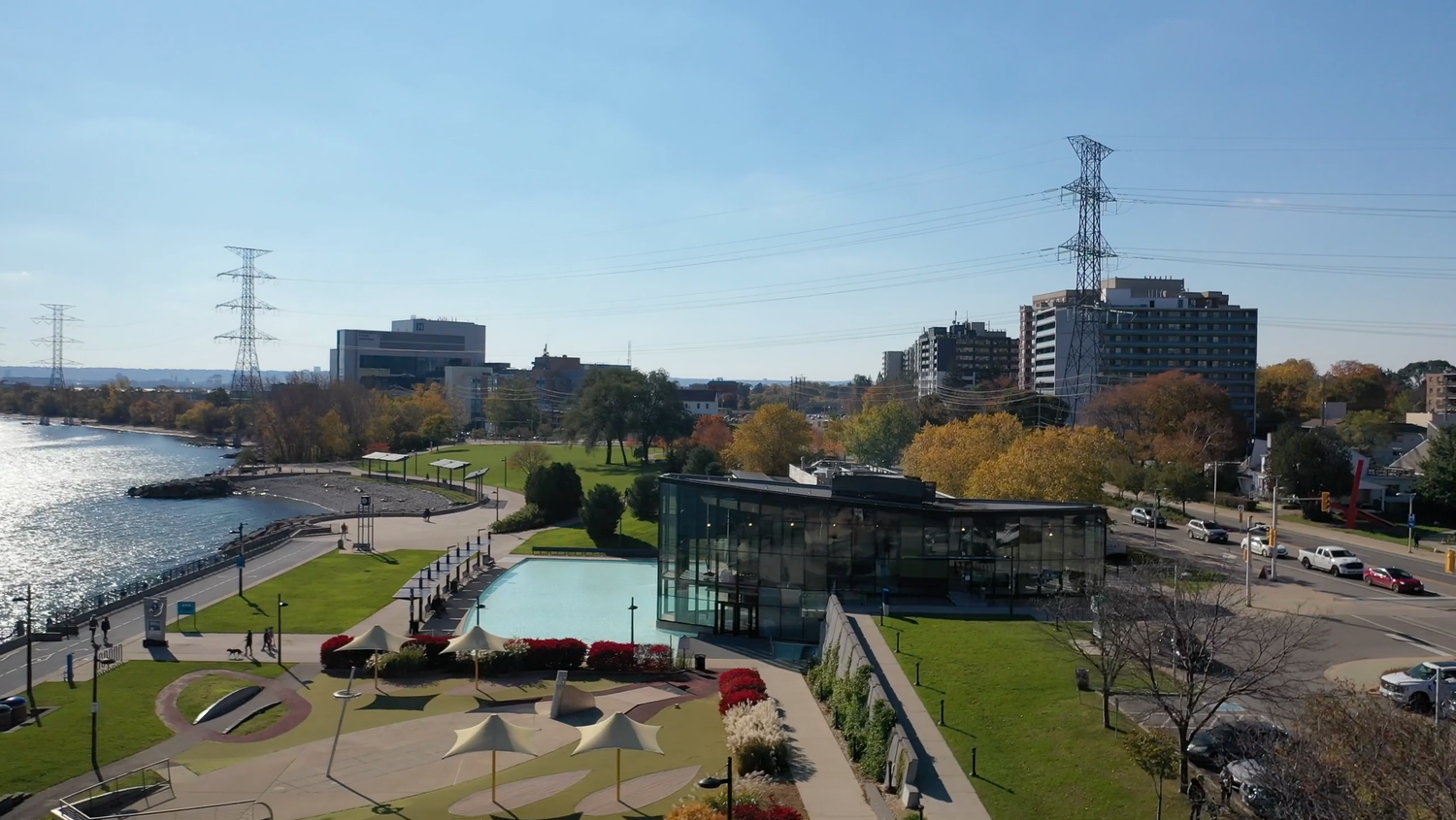 Aerial view of a urban waterfront park with modern buildings, power lines, and colorful autumn trees on a sunny day.