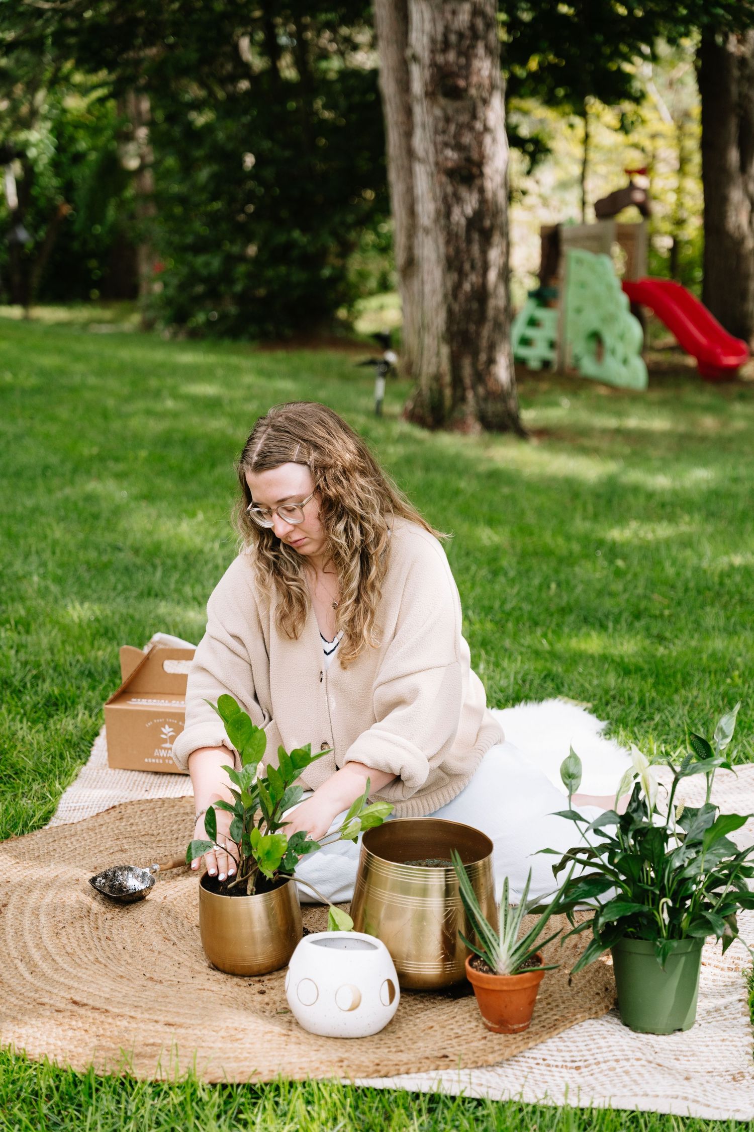 Someone sits on a round woven rug outdoors arranging potted plants and gardening supplies on a sunny day.