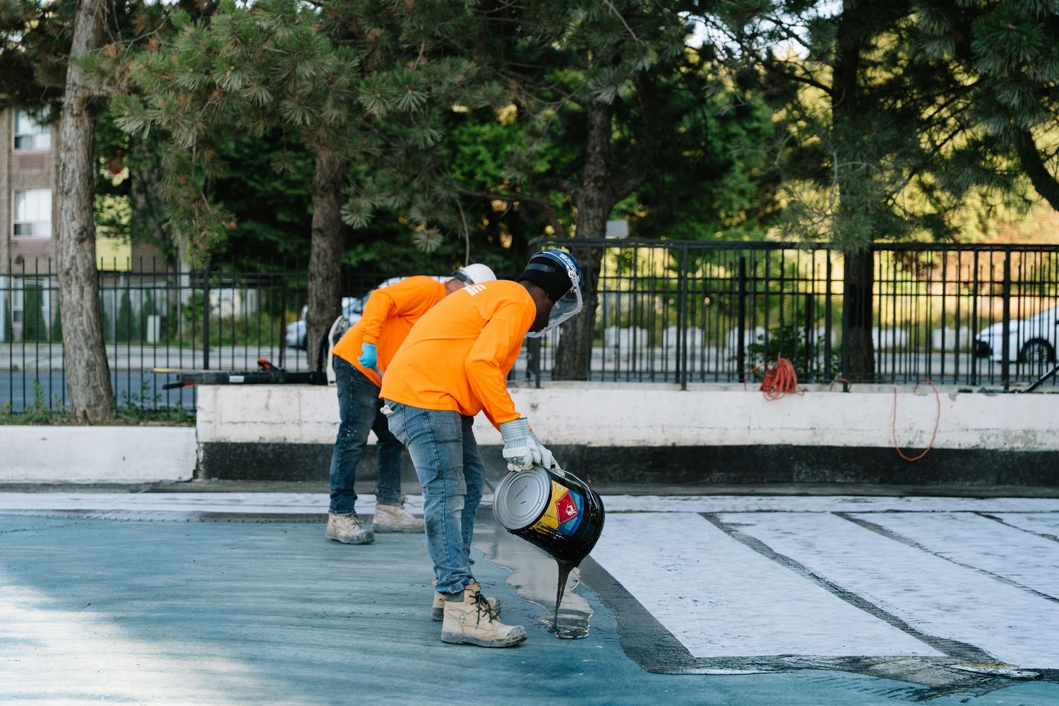 Workers in orange safety gear perform maintenance work on a paved outdoor surface near trees and fencing.