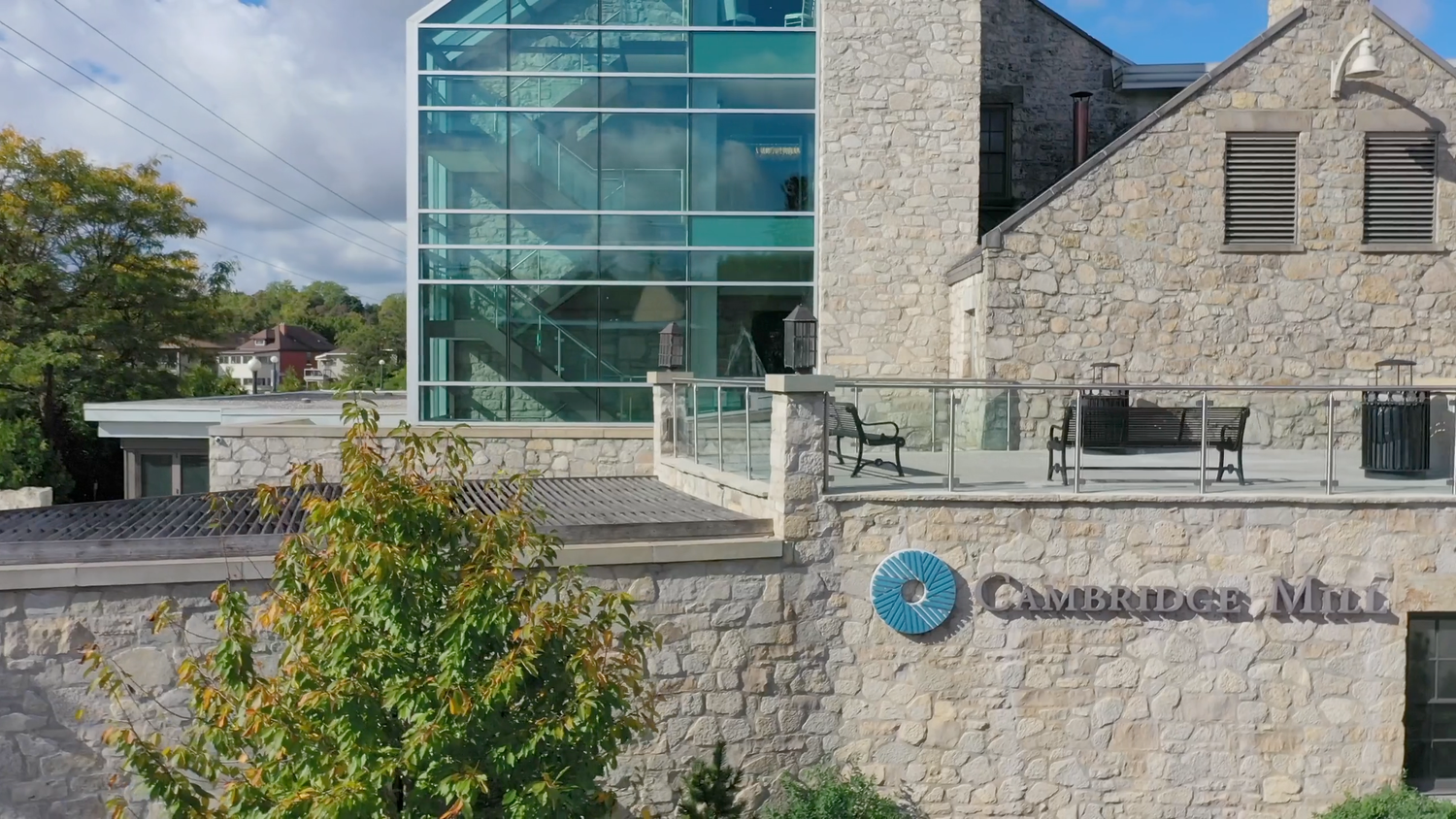 Cambridge Mill building exterior featuring modern glass architecture and historic stone facade with signage, surrounded by lush greenery.
