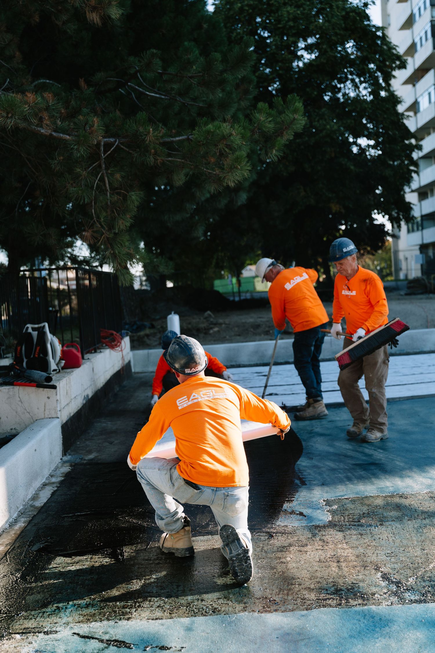 Workers in orange safety vests apply sealant to a concrete surface in sequential action shots.