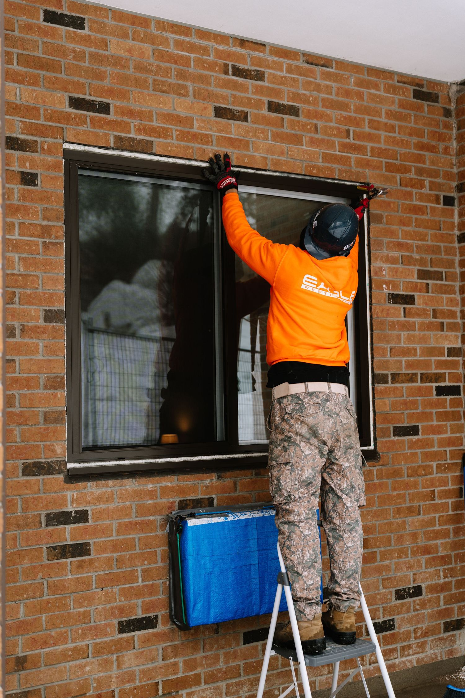 Maintenance worker in orange safety gear works on exterior windows of brick building from ladder.