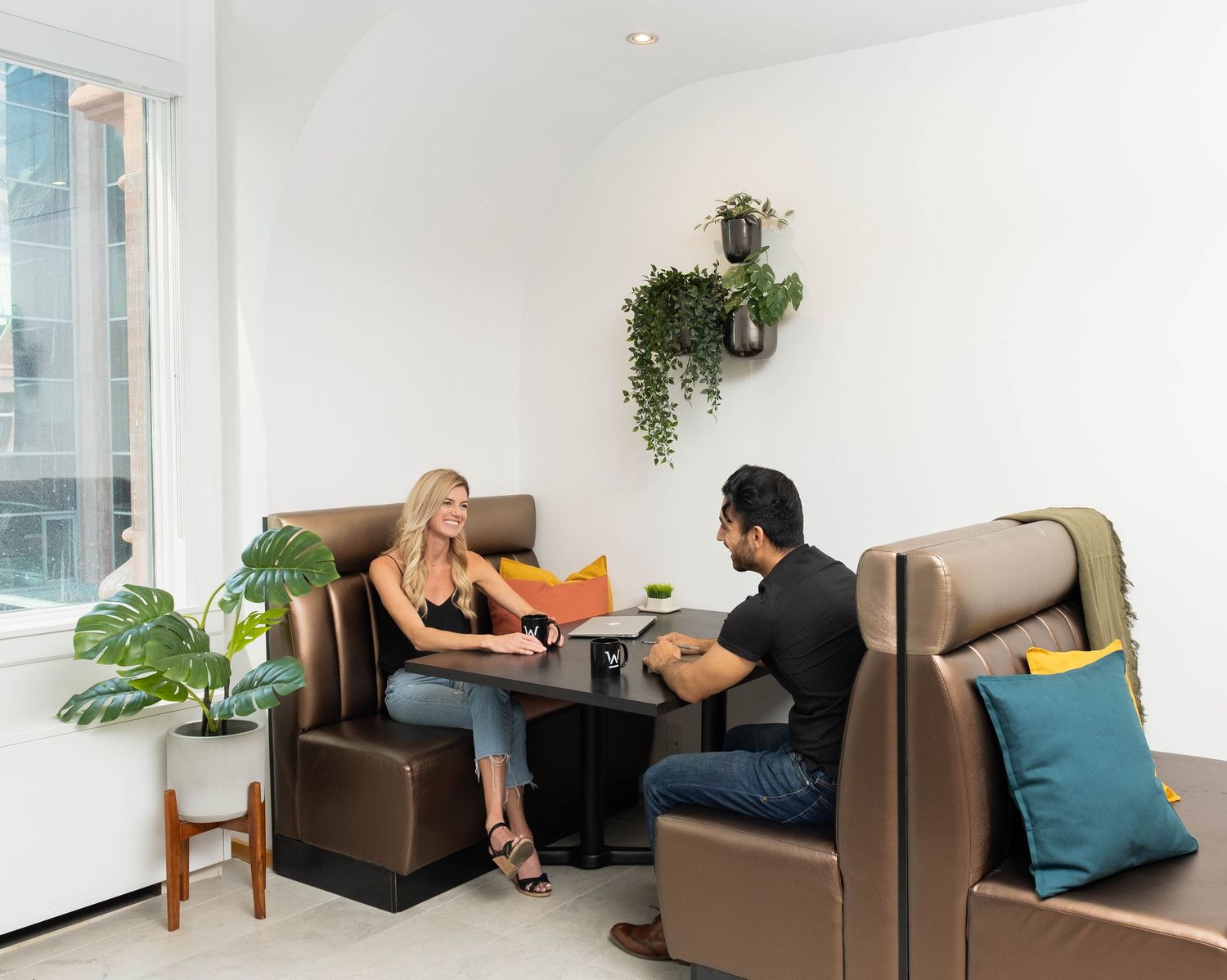 Two people sit at a cozy booth in a modern office space with brown seating and decorative plants.