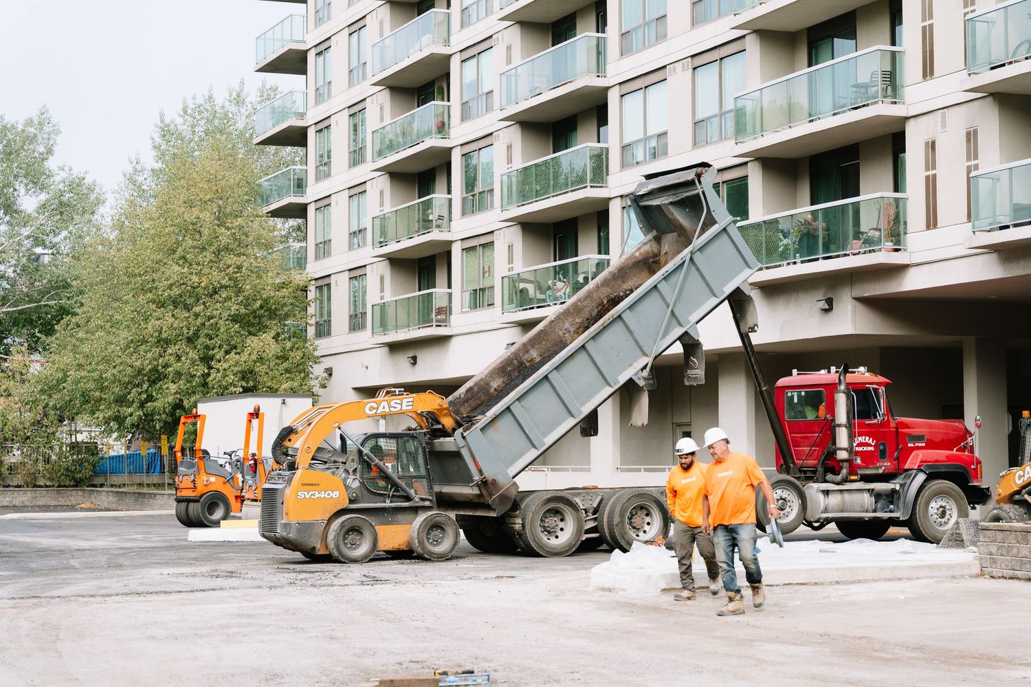Dump trucks and construction equipment working outside a modern apartment building.