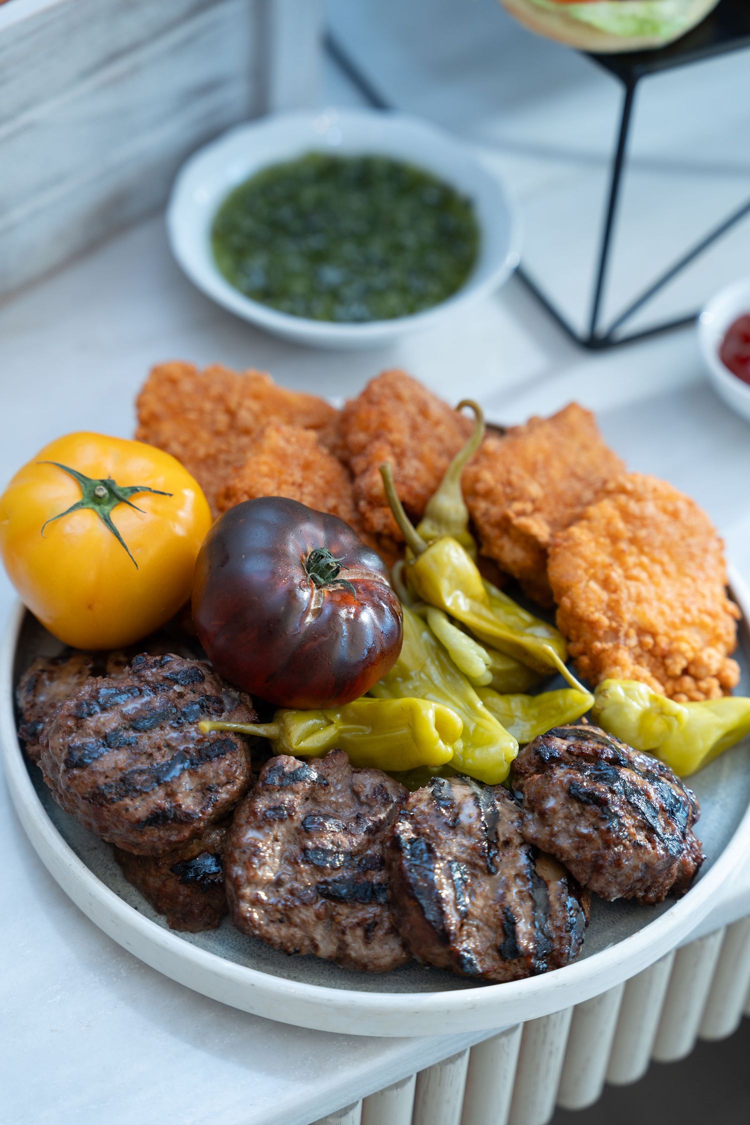 A plate of grilled burger patties served with fried chicken, pepperoncini peppers, heirloom tomatoes and green sauce.