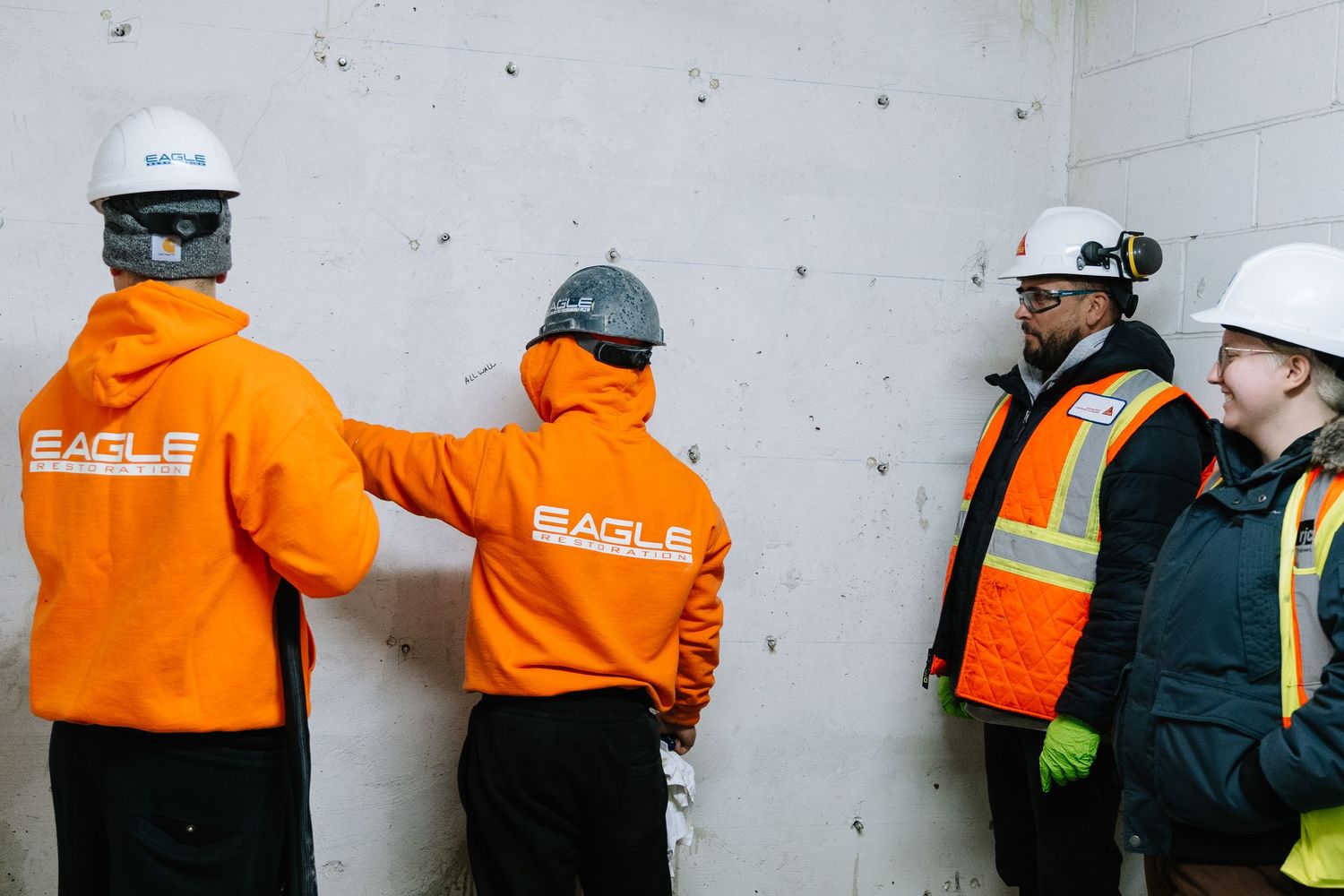 Workers in orange safety uniforms and protective gear inspect a concrete wall during construction.