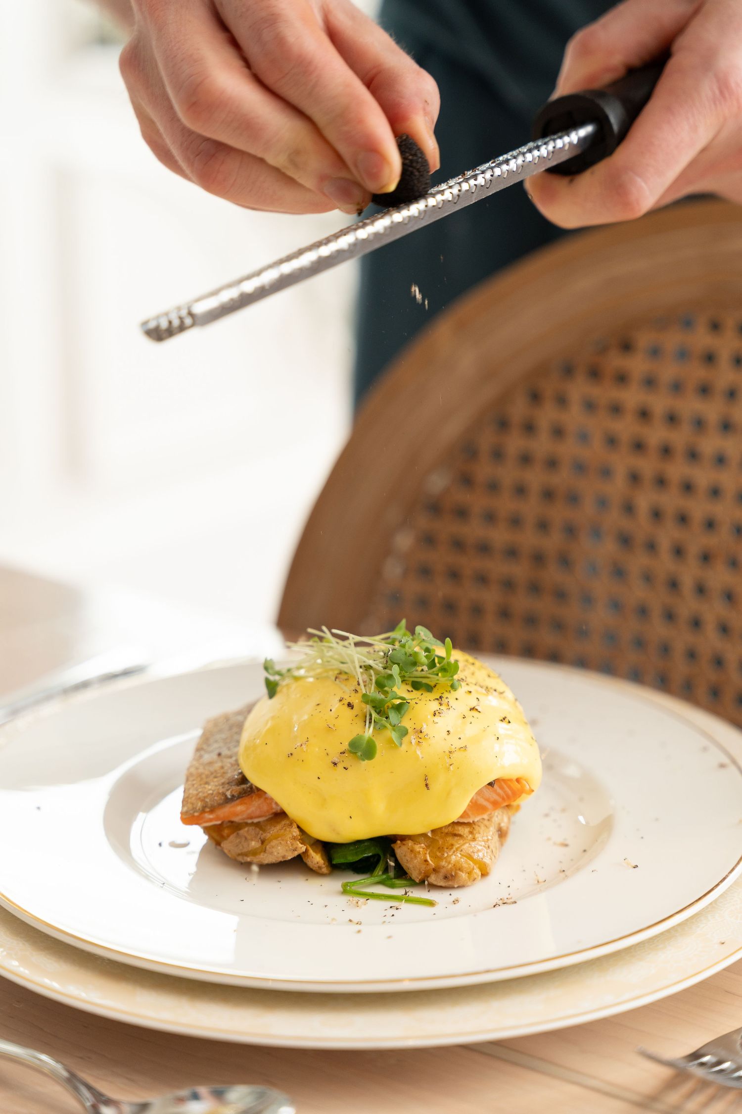 Eggs Benedict with hollandaise sauce and microgreens being seasoned on white plate.
