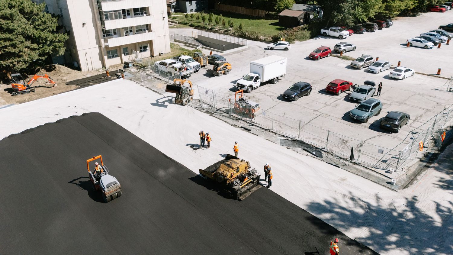 Birds eye view of construction workers resurfacing a parking lot next to parked cars and buildings.