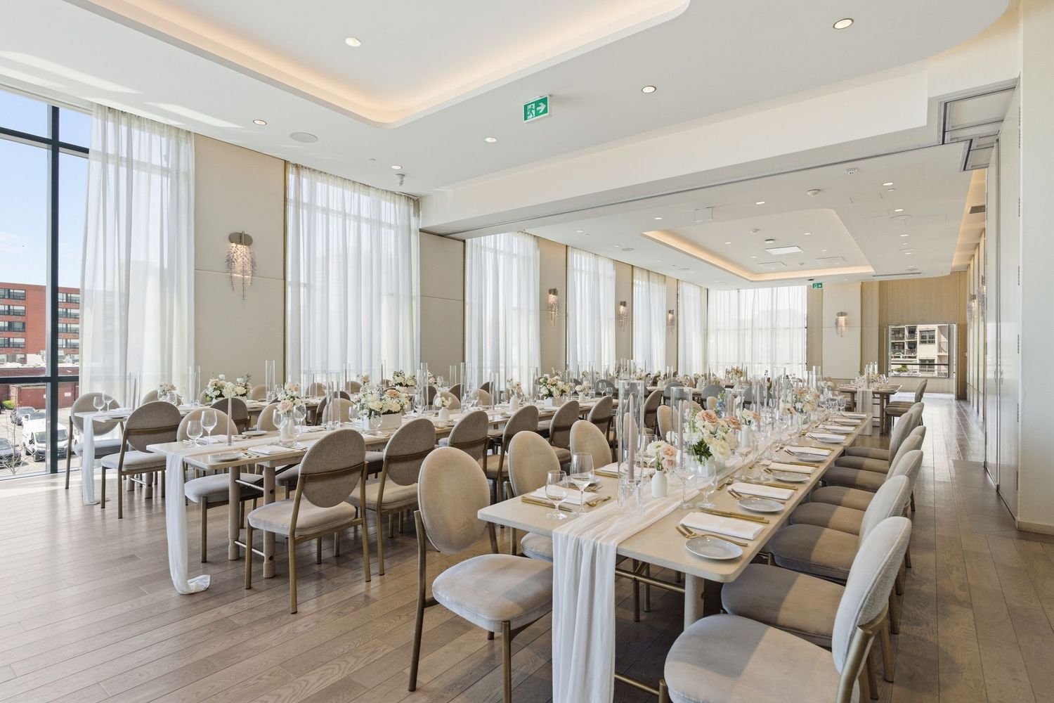 Elegant restaurant interior with cream colored chairs and tables arranged in a long bright dining room with recessed lighting.