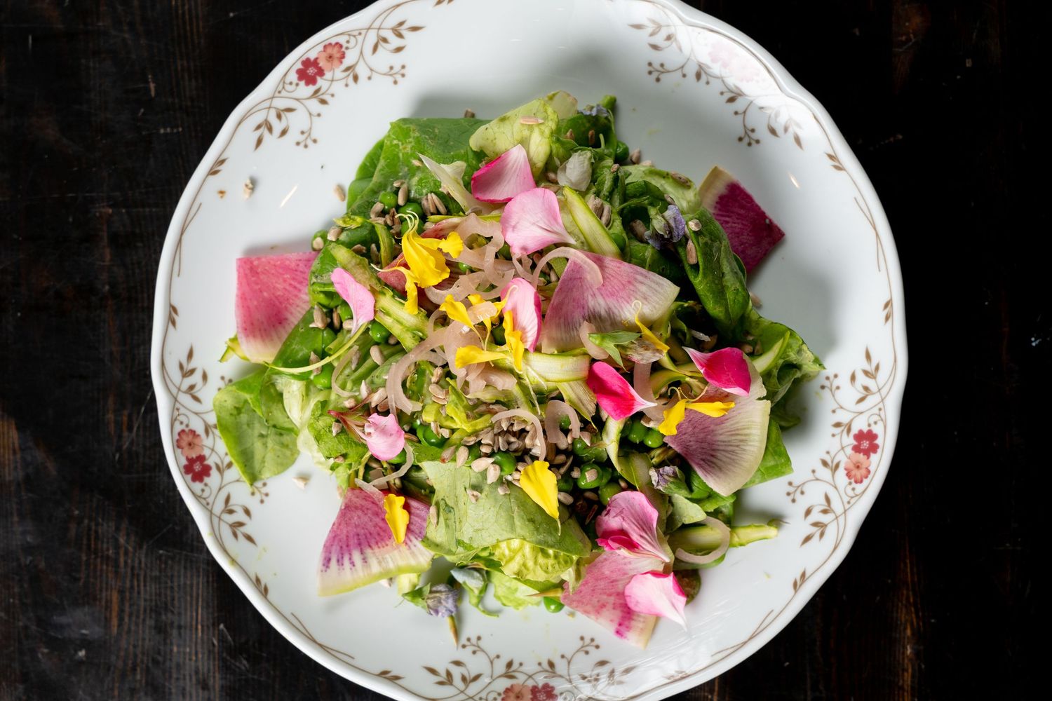 Elegant white floral plate with fresh green salad topped with edible flowers, microgreens, and colorful radish slices.