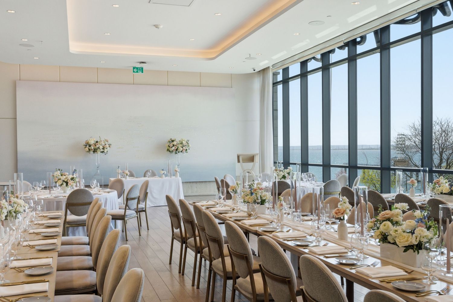 Elegant wedding reception venue with long dining tables, floor-to-ceiling windows, and white floral centerpieces overlooking scenic views.