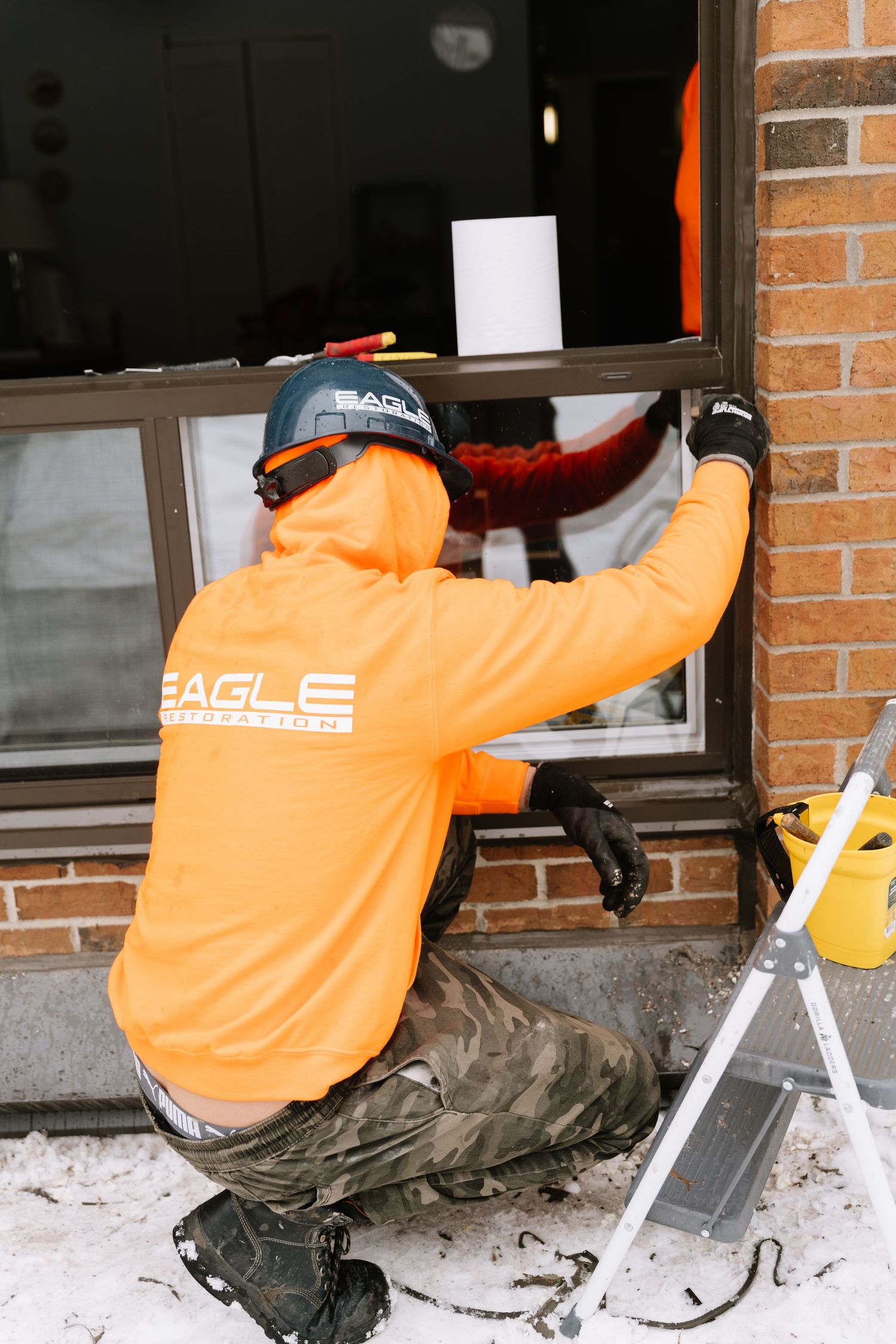 Maintenance worker in orange safety vest performs repairs on snowy window ledge.