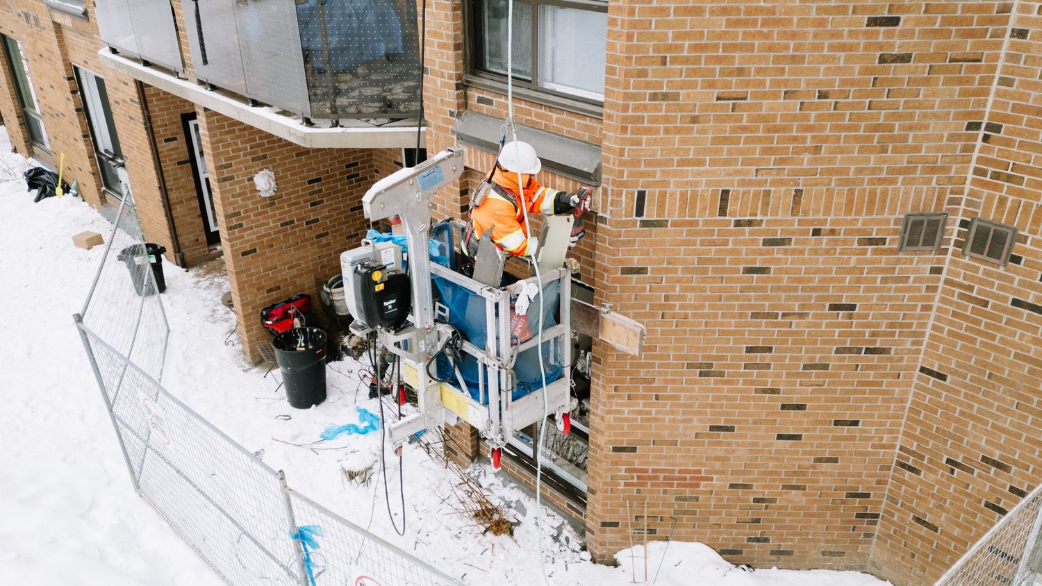 Two dogs play in deep snow outside a brick building during winter, with one dog leaping through the powder.