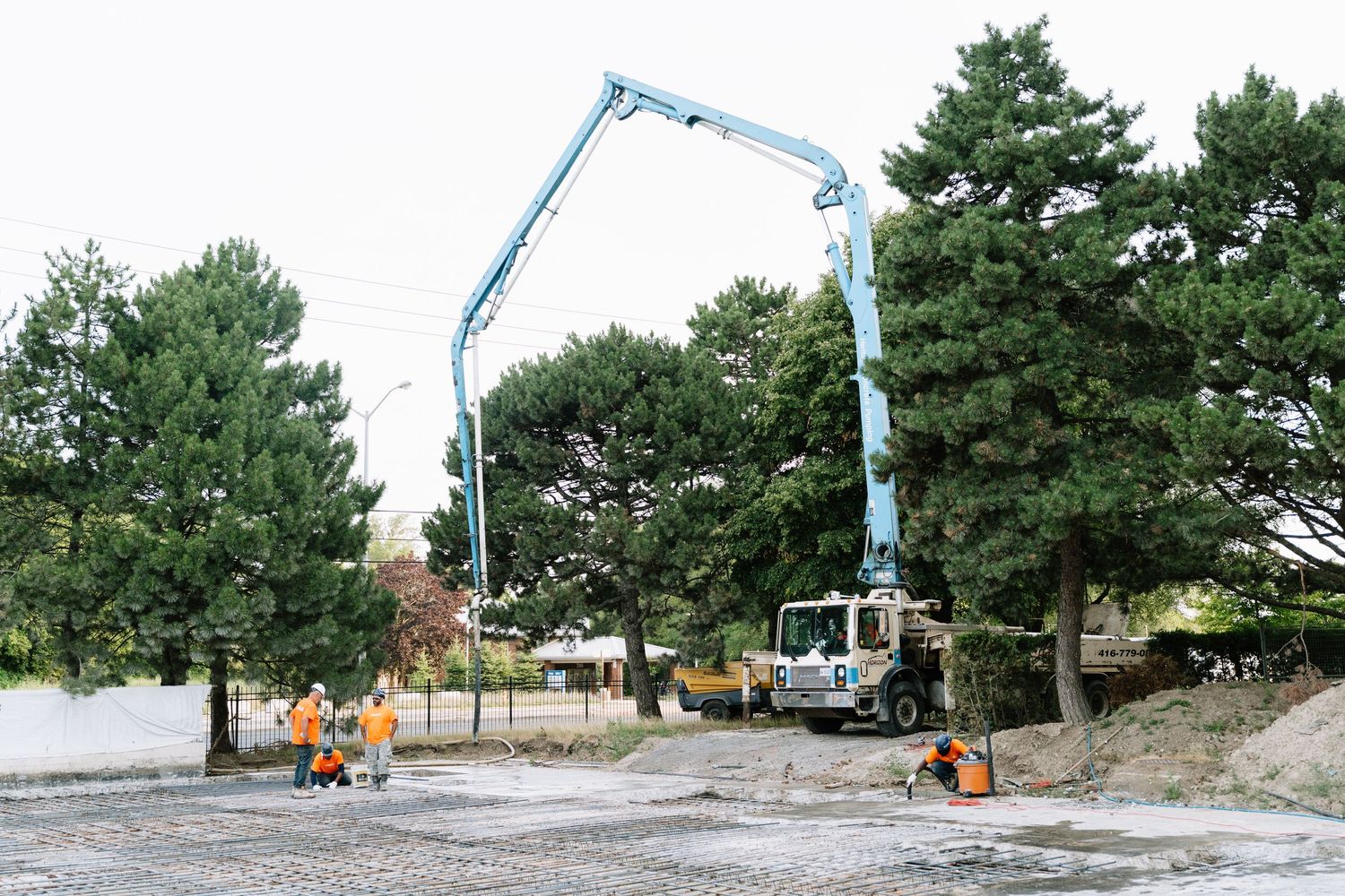 A concrete pump truck with extended blue boom arm operates near tall pine trees at a construction site.