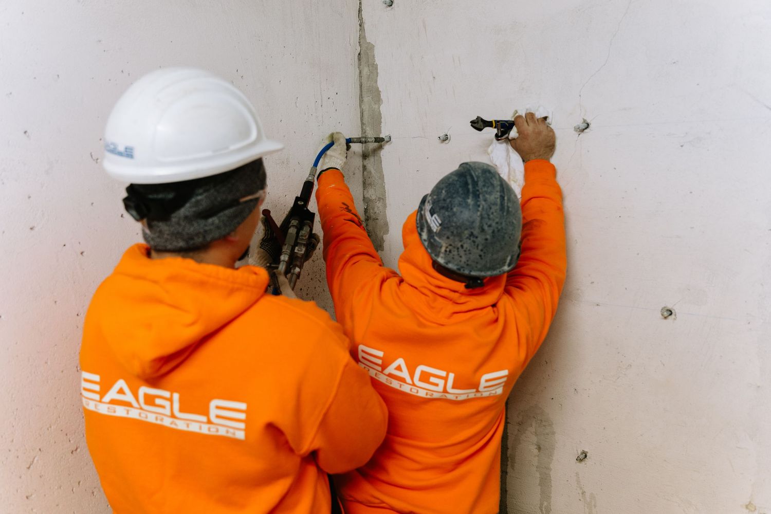Construction workers in orange safety shirts install equipment on a concrete wall.