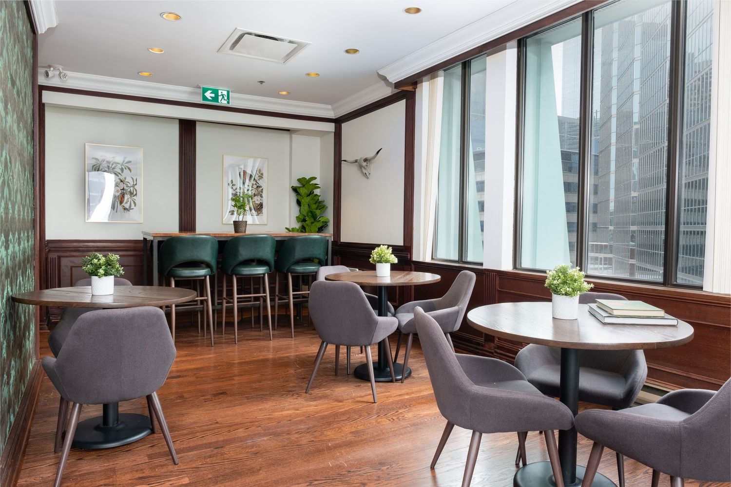 Modern office lounge area with wooden floors, gray chairs, and large windows offering natural light and city views.
