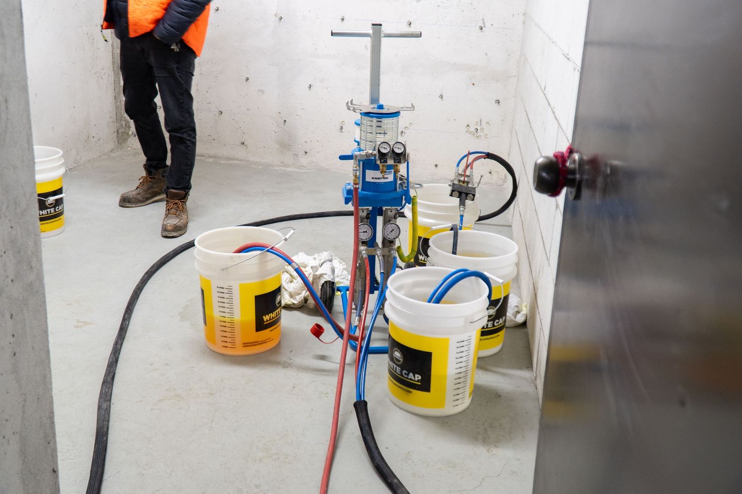 Construction site equipment showing a blue vertical drill setup with yellow chemical containers and black hoses on concrete floor.