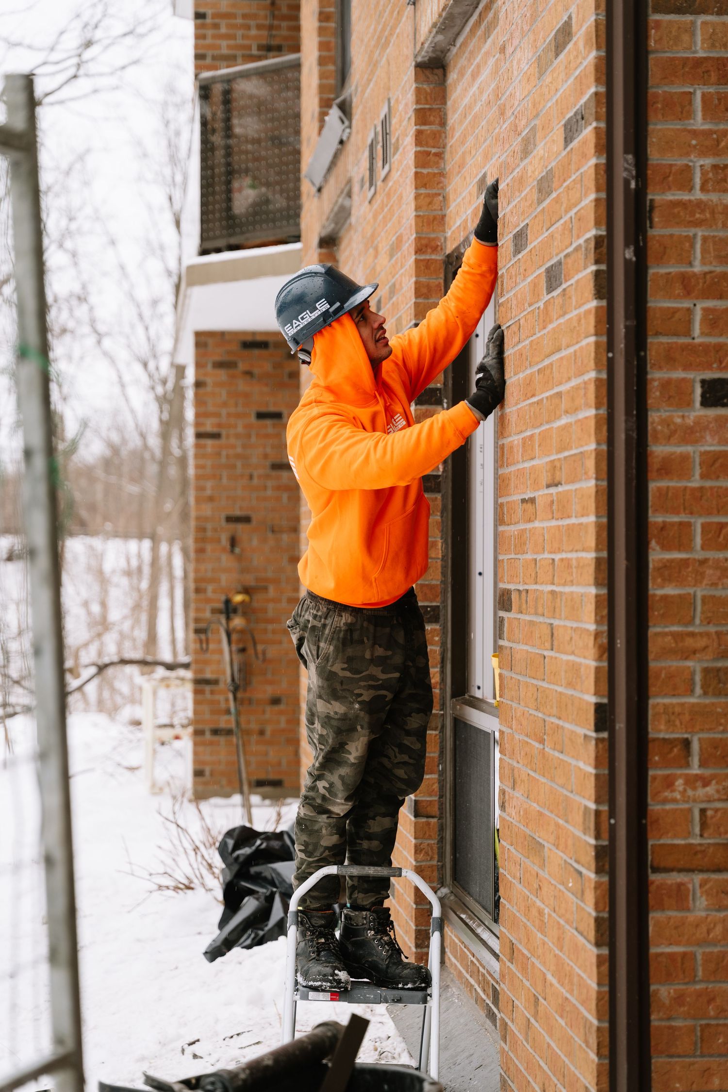 Worker in high-visibility orange jacket services windows on brick building during winter.
