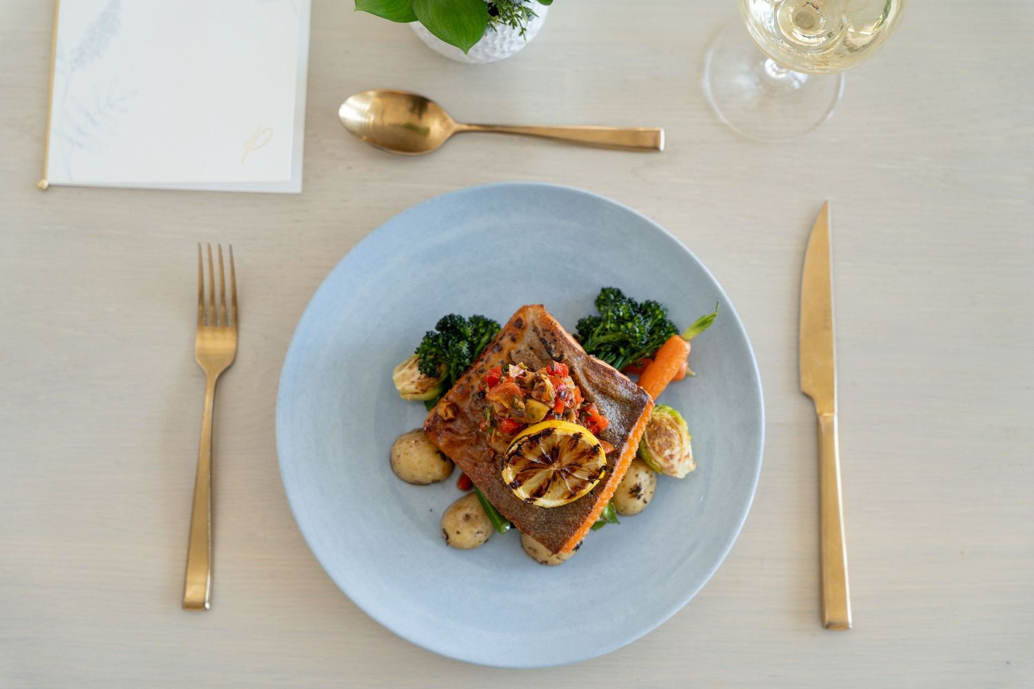 White plates with gourmet meals arranged on a light colored dining table with gold utensils.