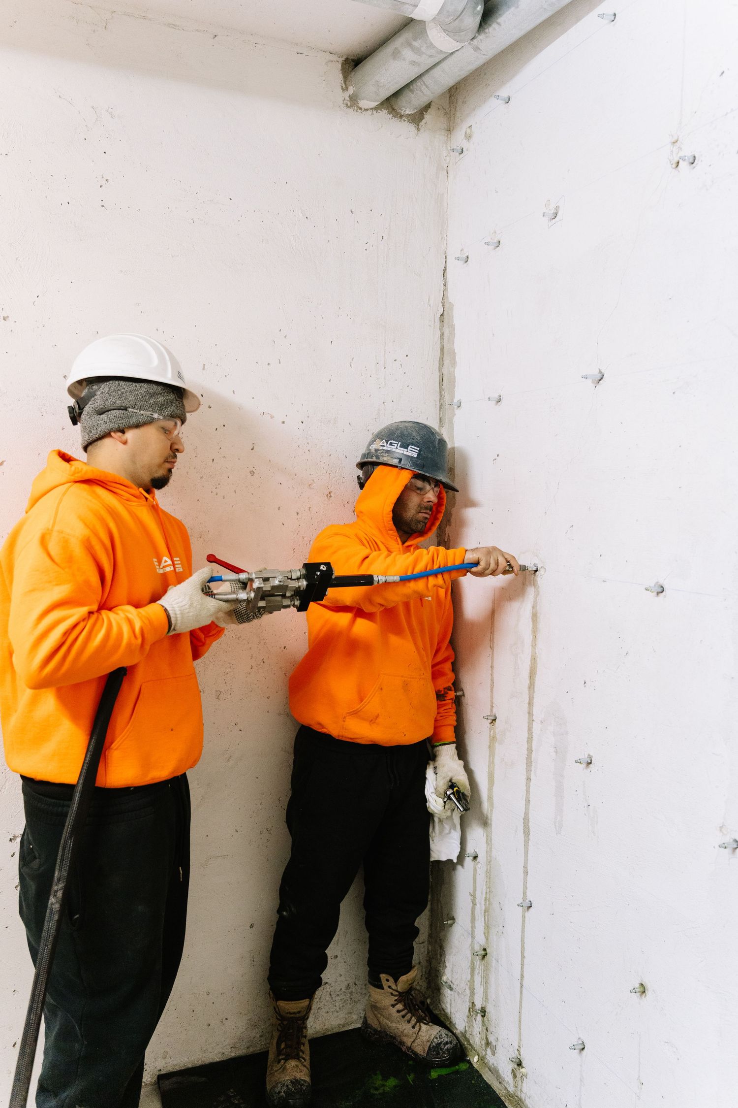 Construction workers in orange safety shirts inspect and repair walls in a white interior space.
