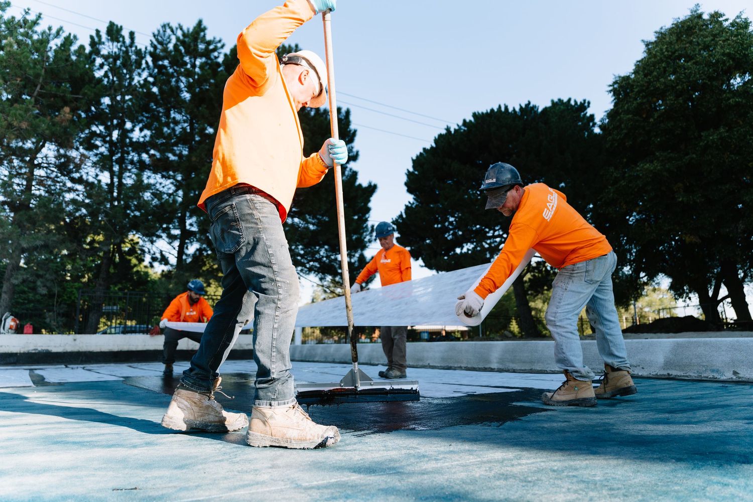 Construction workers in bright orange safety gear smoothing wet concrete at an outdoor worksite.