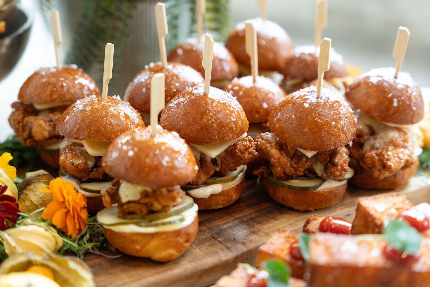 Row of gourmet mini sliders with brioche buns and fresh toppings served on wooden boards at a catering event.