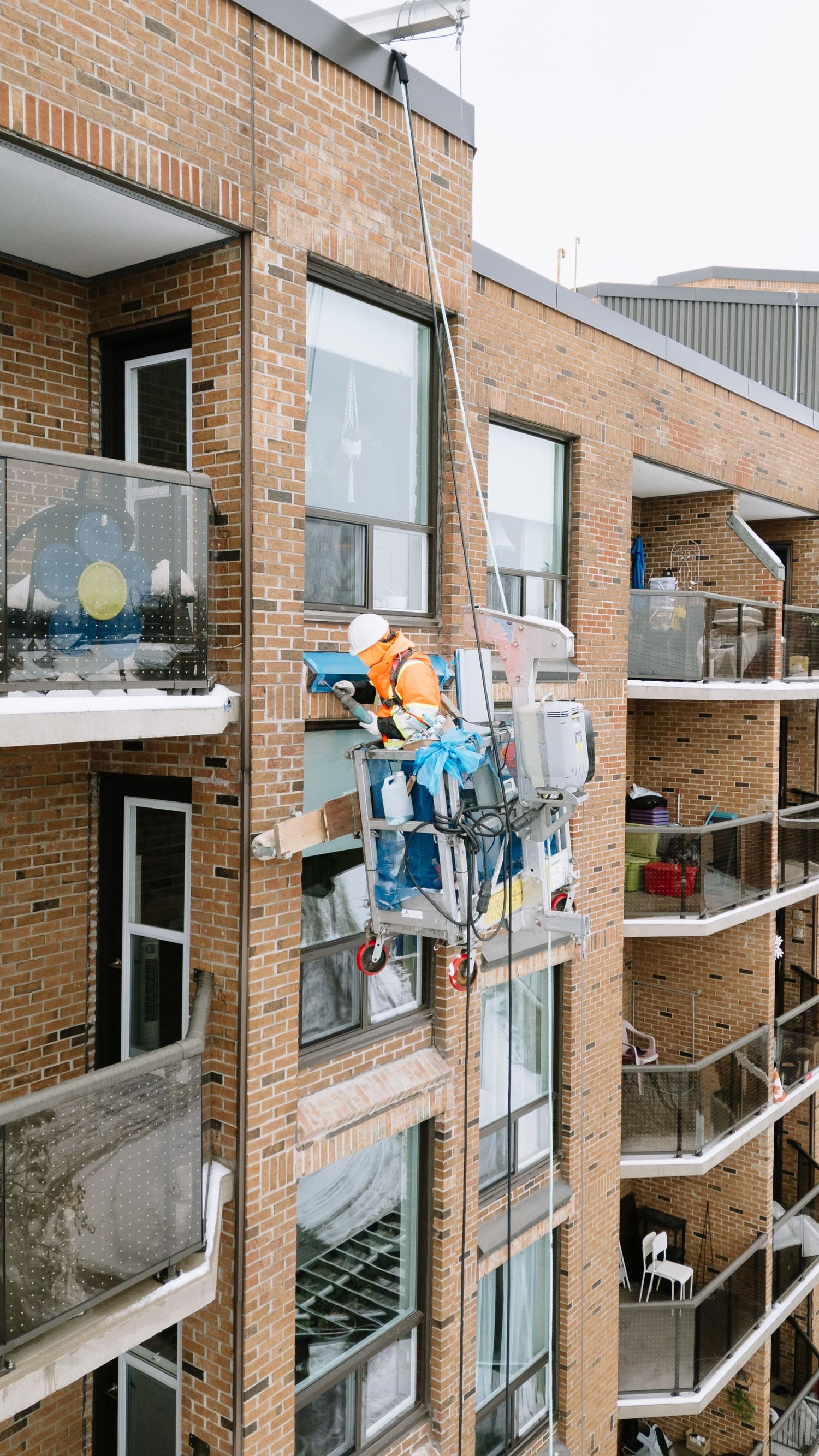 Window cleaner in bright orange safety gear works on exterior windows of brick apartment building.